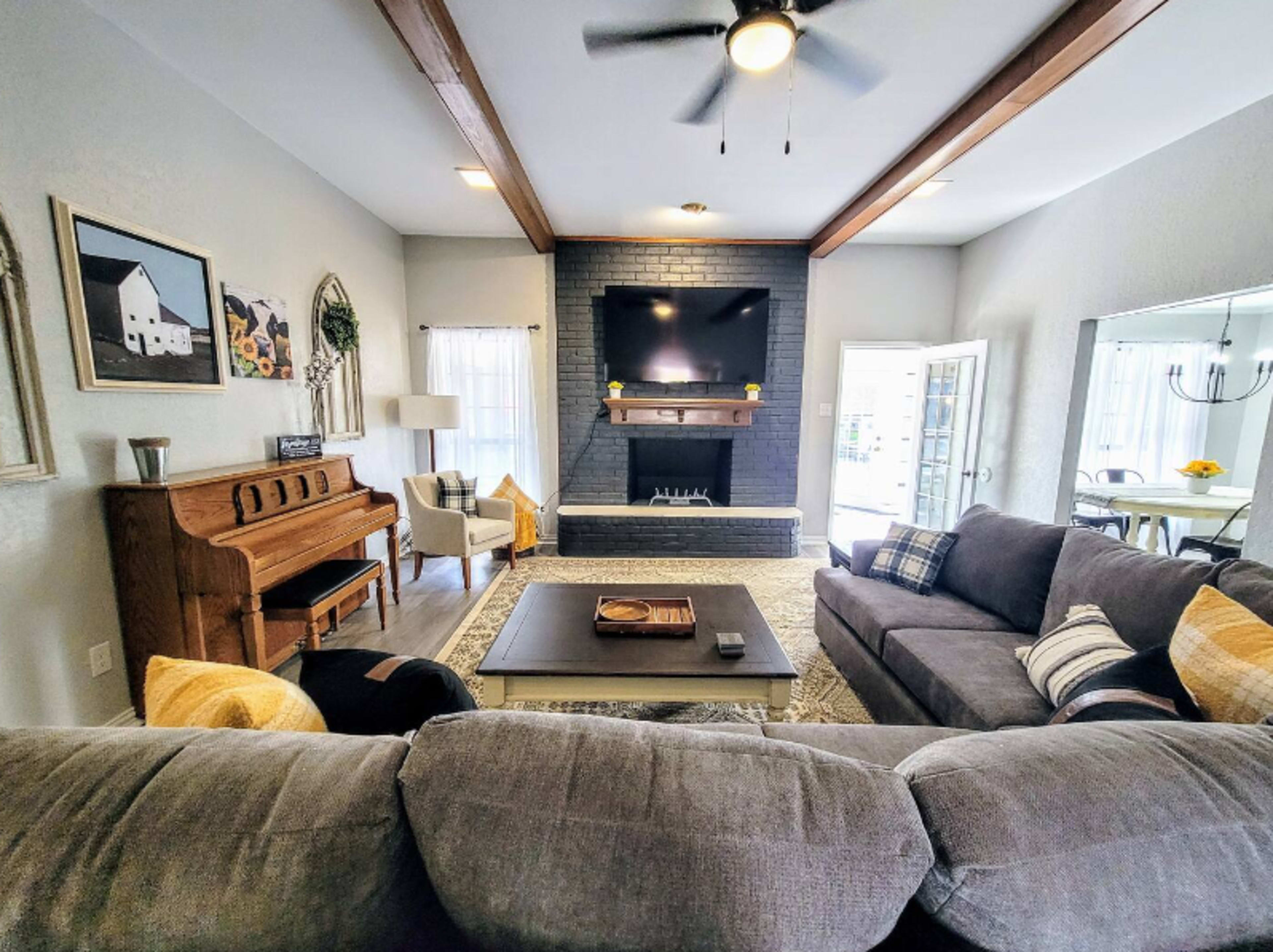 The living room features a sofa and armchairs positioned around a coffee table, with a piano against one wall and a fireplace with a mounted television above it.