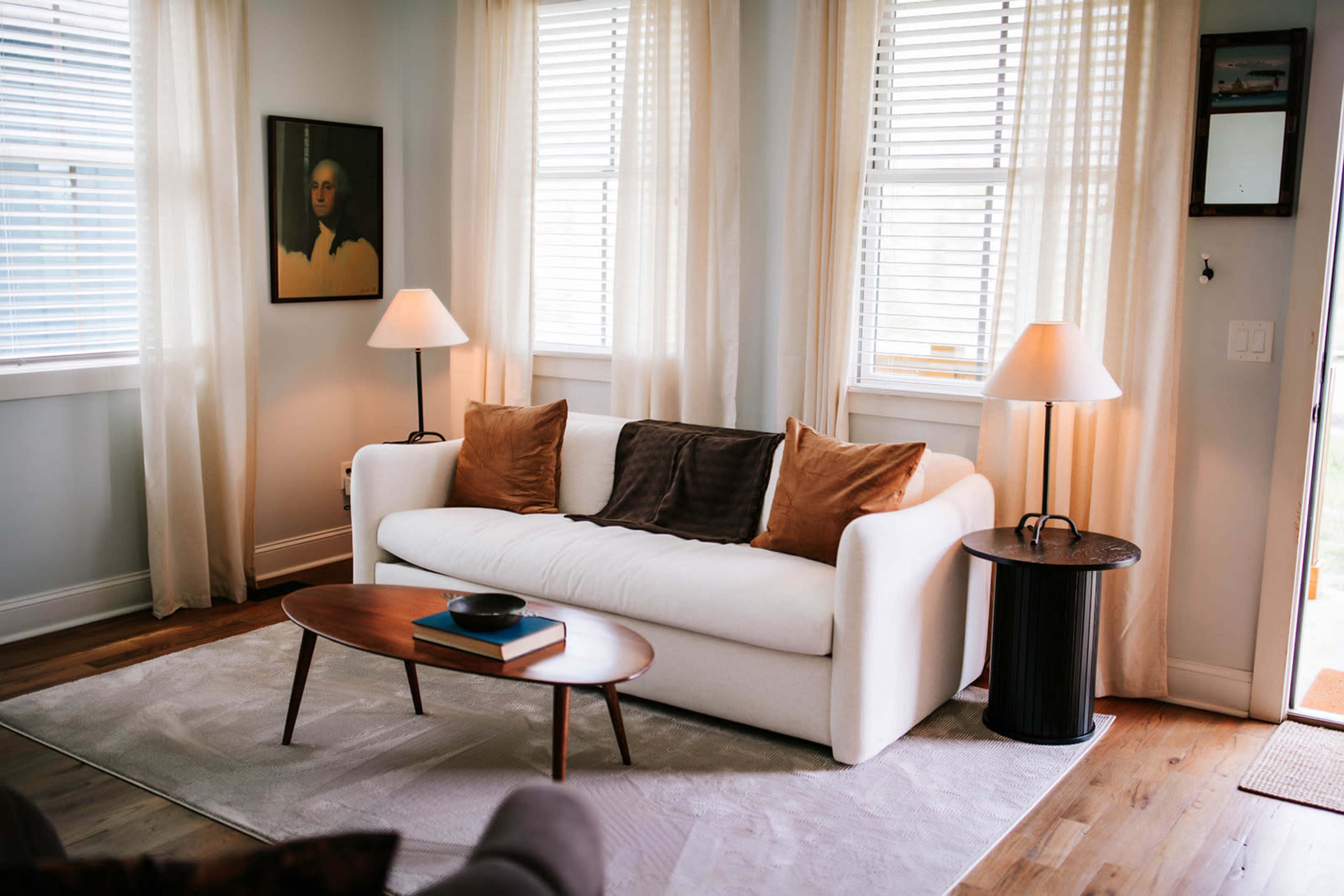A light-filled living room features a white sofa with brown cushions, an oval wooden coffee table, and two lamps beside large windows.