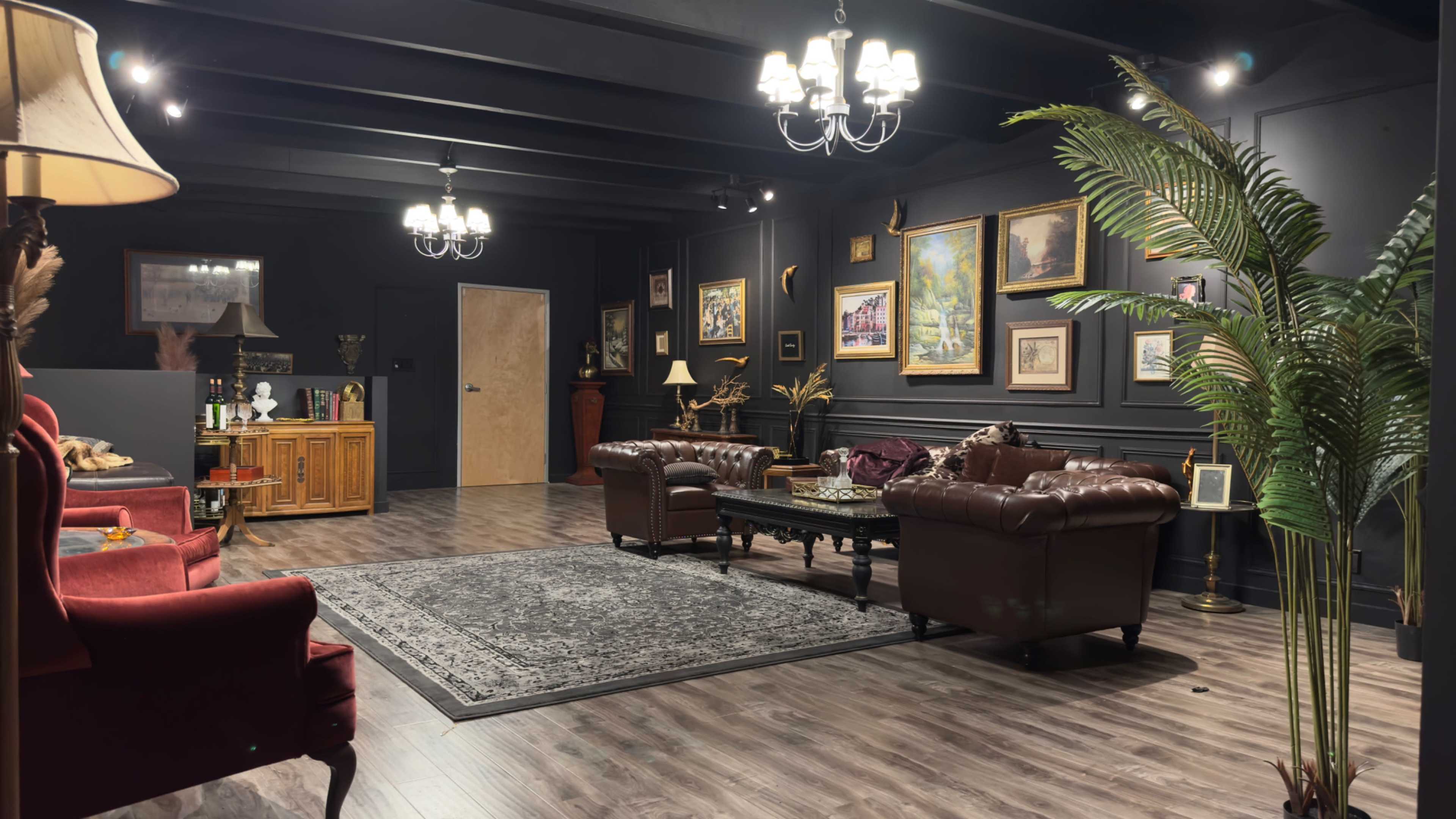 The image shows a darkly decorated room with leather sofas, wooden furniture, framed artwork, and a large area rug, illuminated by ceiling lights and chandeliers.