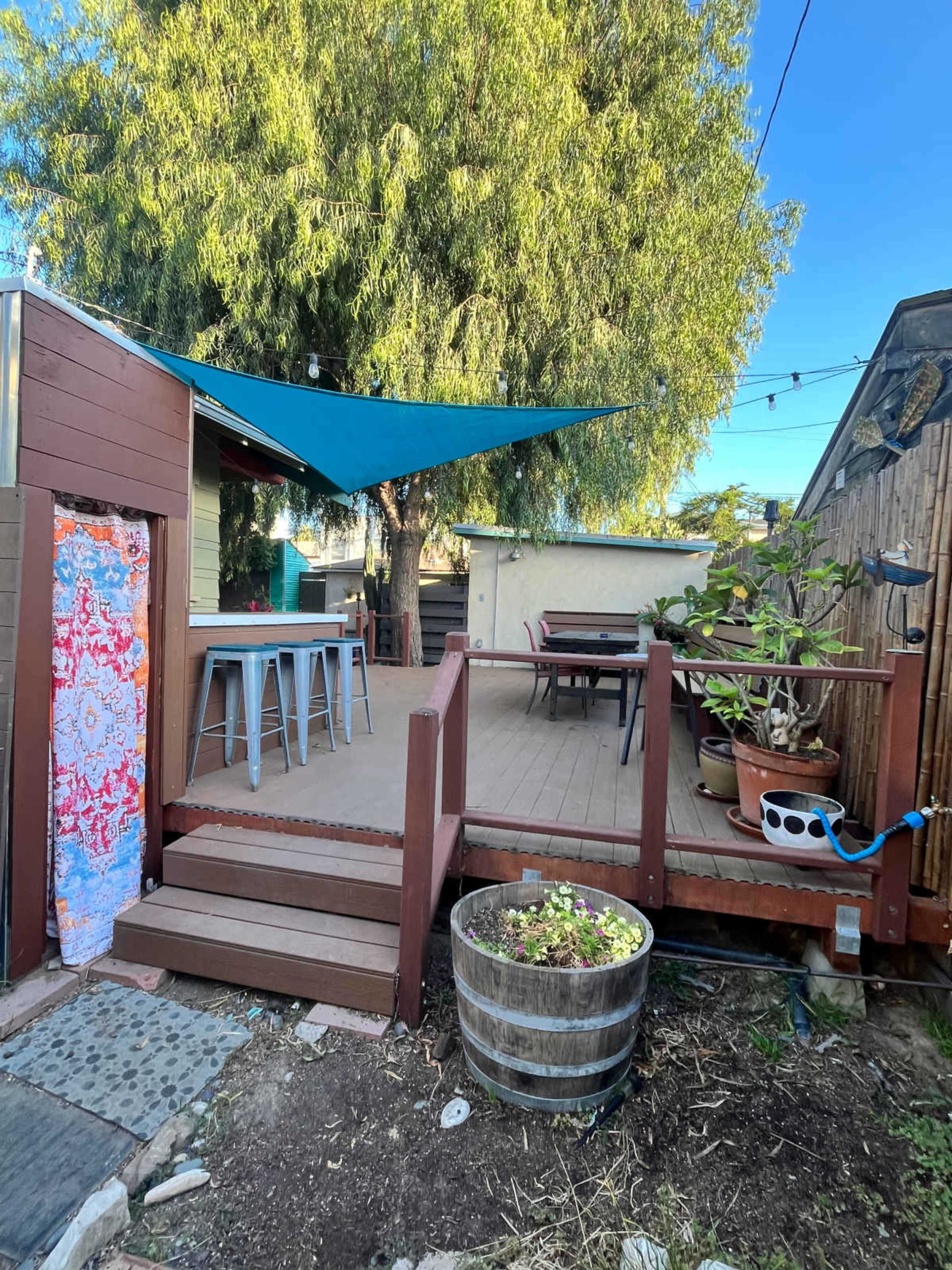 The image shows a wooden deck area with a blue shade sail, a table and chairs, and potted plants surrounding it.
