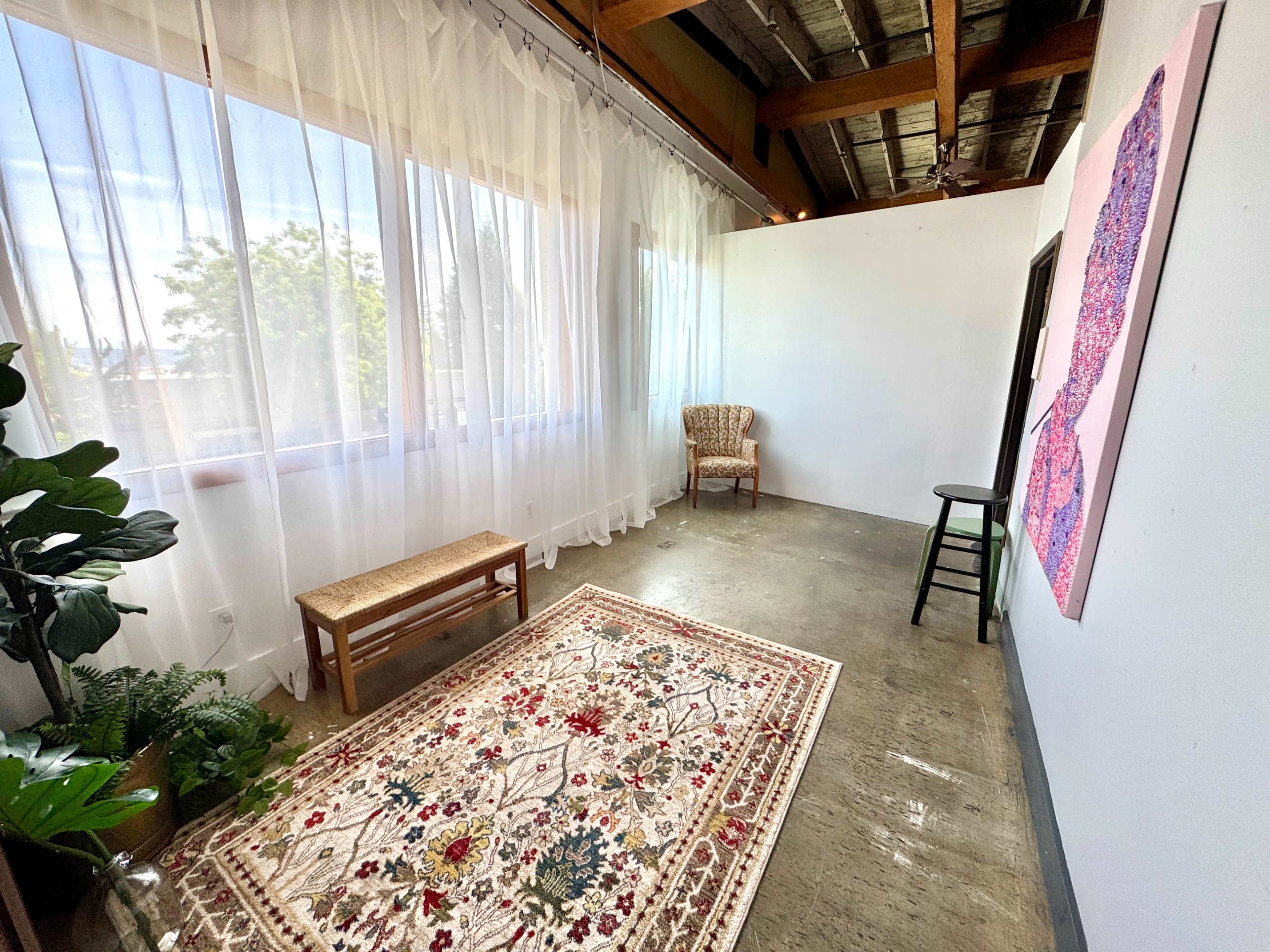 The image shows a bright room with large windows, a patterned rug on the floor, and minimal furnishings including a chair and a bench.