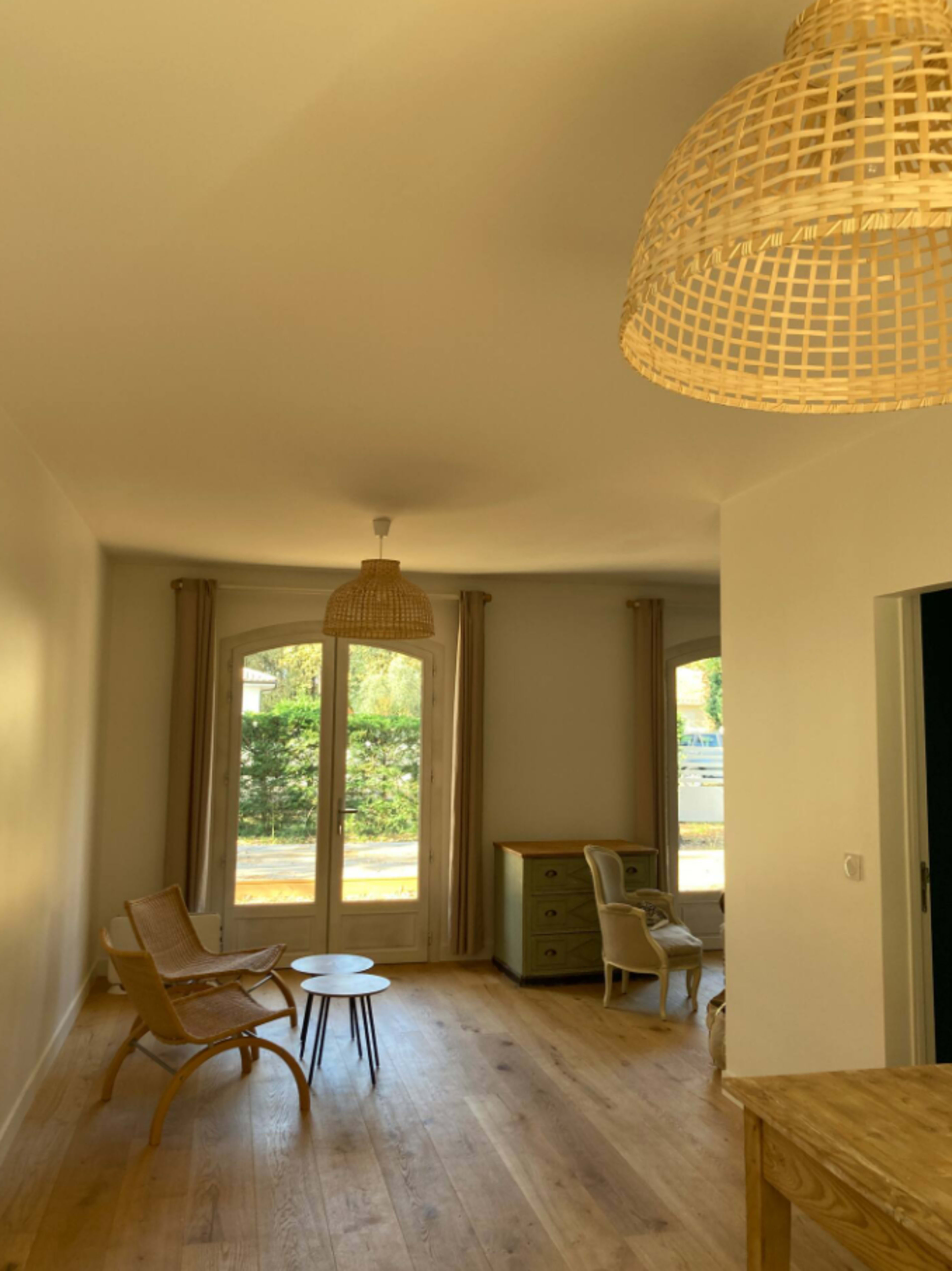 A bright room features wicker pendant lights, wooden floors, and a small table surrounded by chairs near large windows.