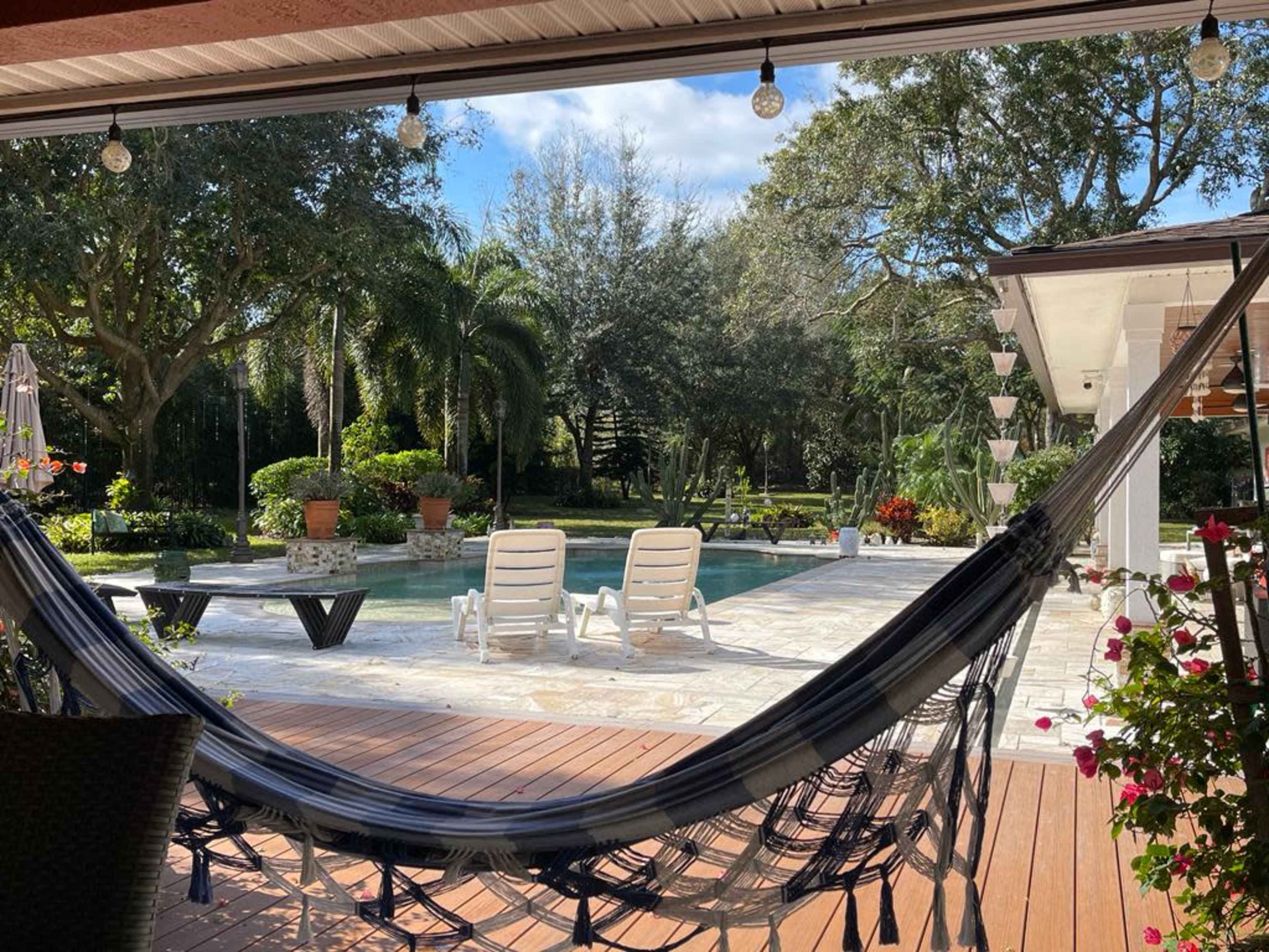 A hammock hangs between posts overlooking a pool surrounded by greenery and lounge chairs.