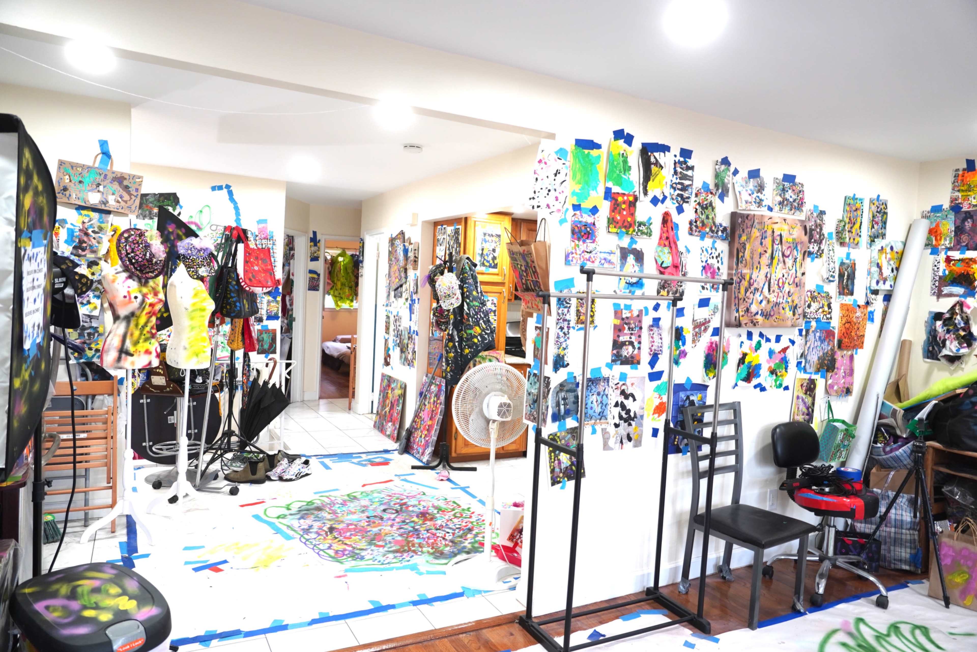 The image shows an art studio filled with colorful paintings and collages hanging on the walls, along with various art supplies and furniture scattered around the space.