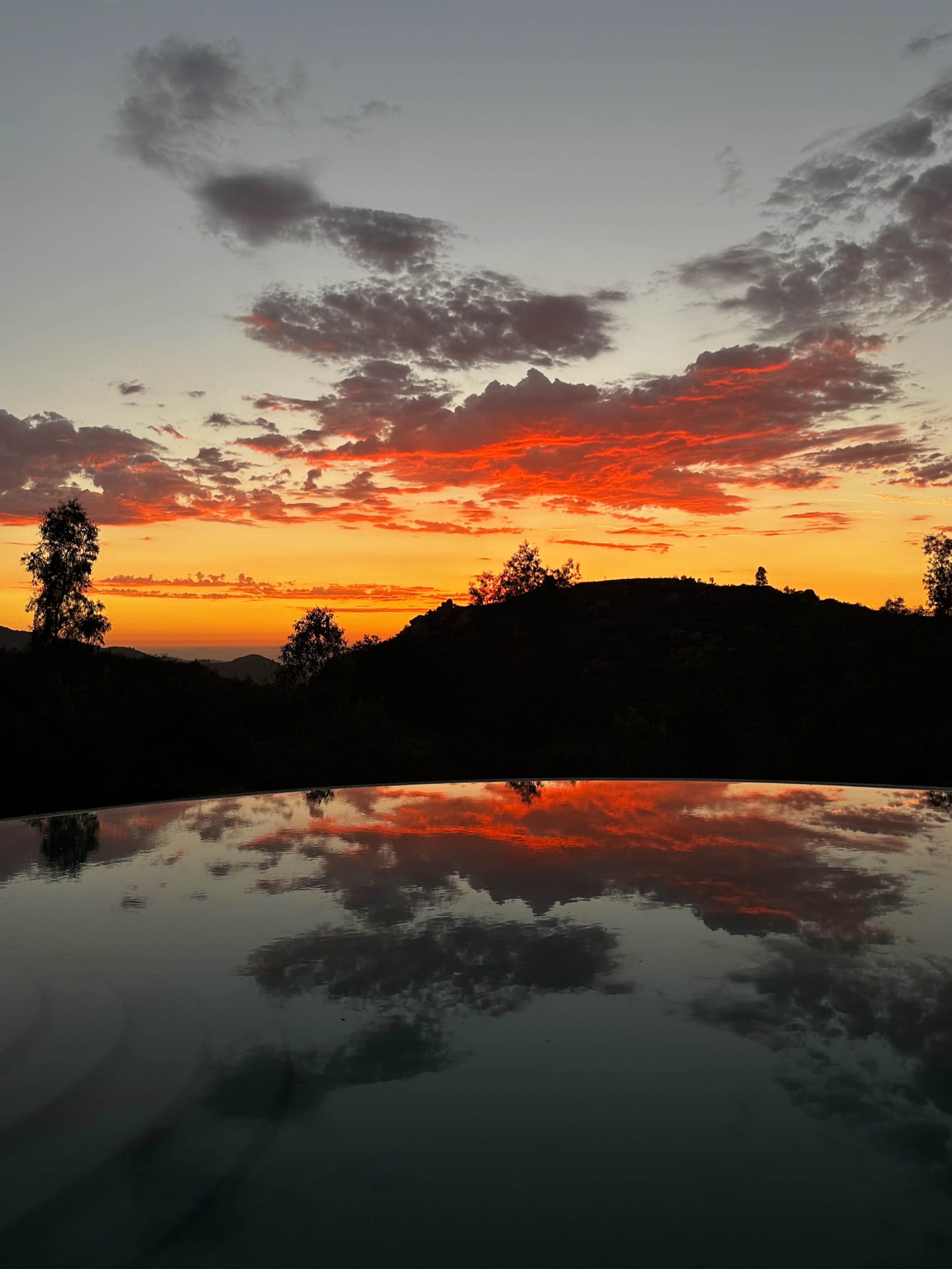 The image shows a sunset with vibrant clouds reflected in the calm water of a pool, overlooking a mountain silhouette.