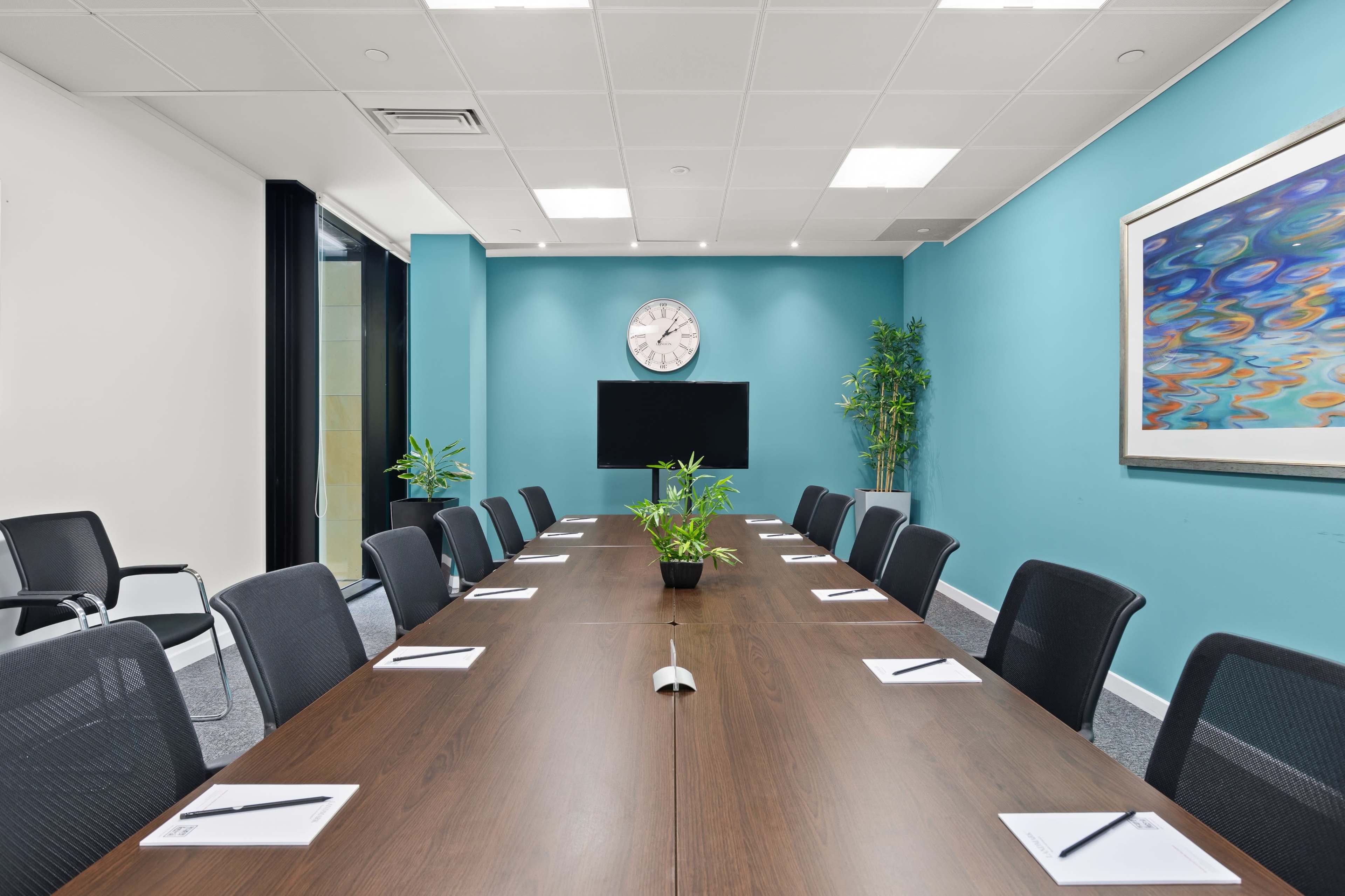 A modern conference room features a long wooden table, black chairs, a clock on the wall, and a television mounted on a turquoise wall.