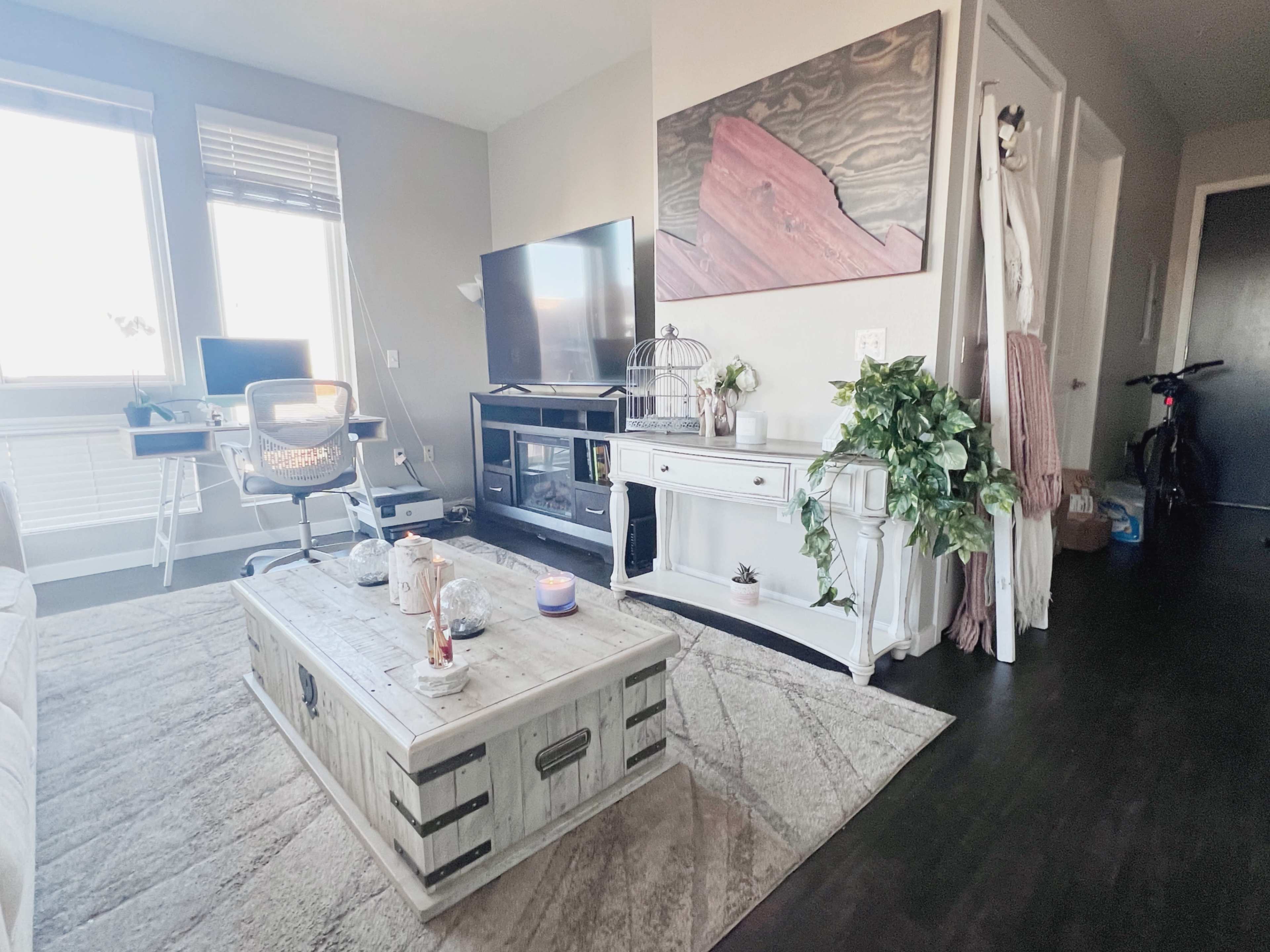 A modern living room with a wooden coffee table, a television on a stand, a desk with a chair, and potted plants.