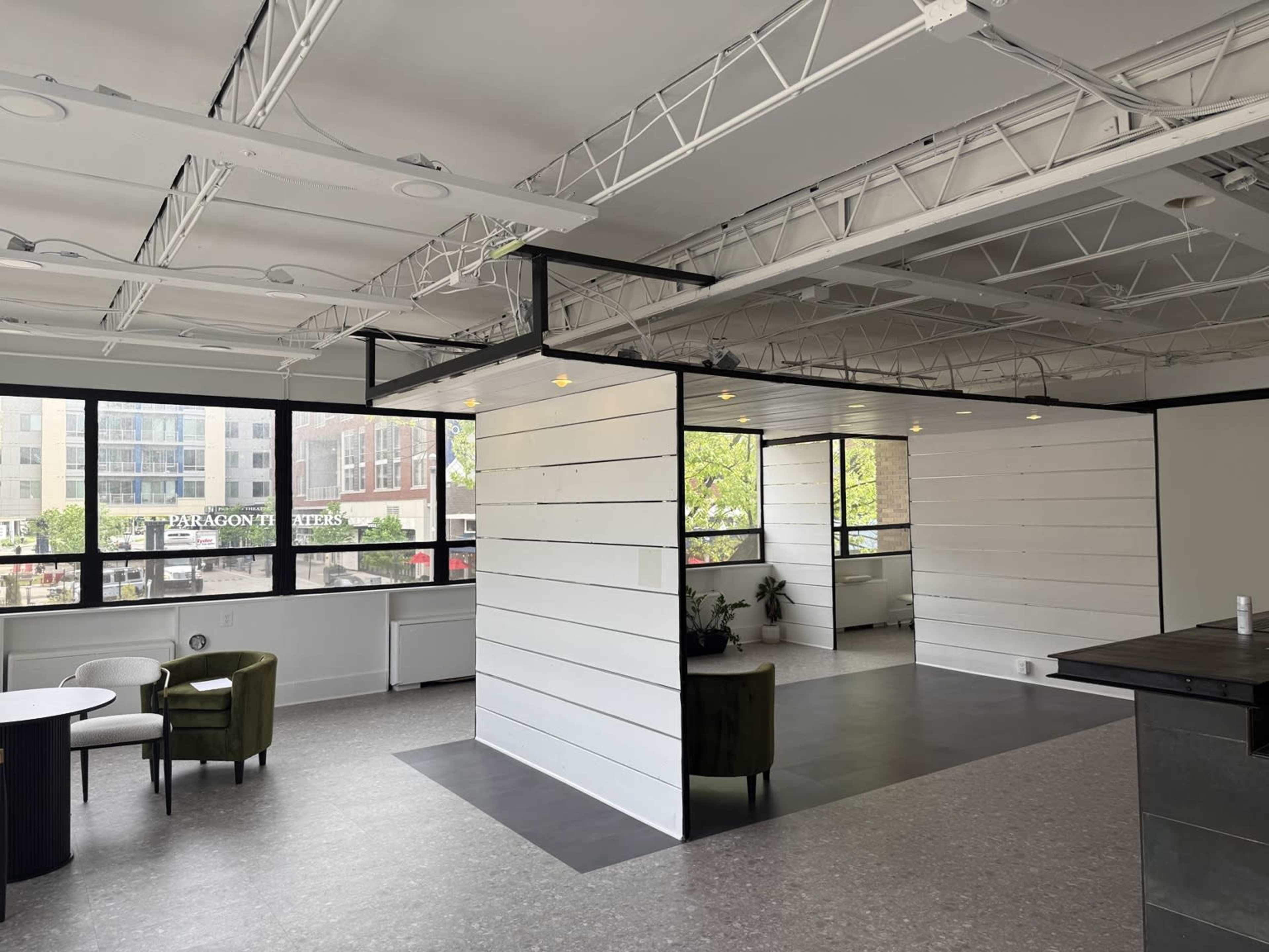 The image shows a modern open space with white wooden paneling, large windows, and seating areas featuring two green chairs and a small round table.