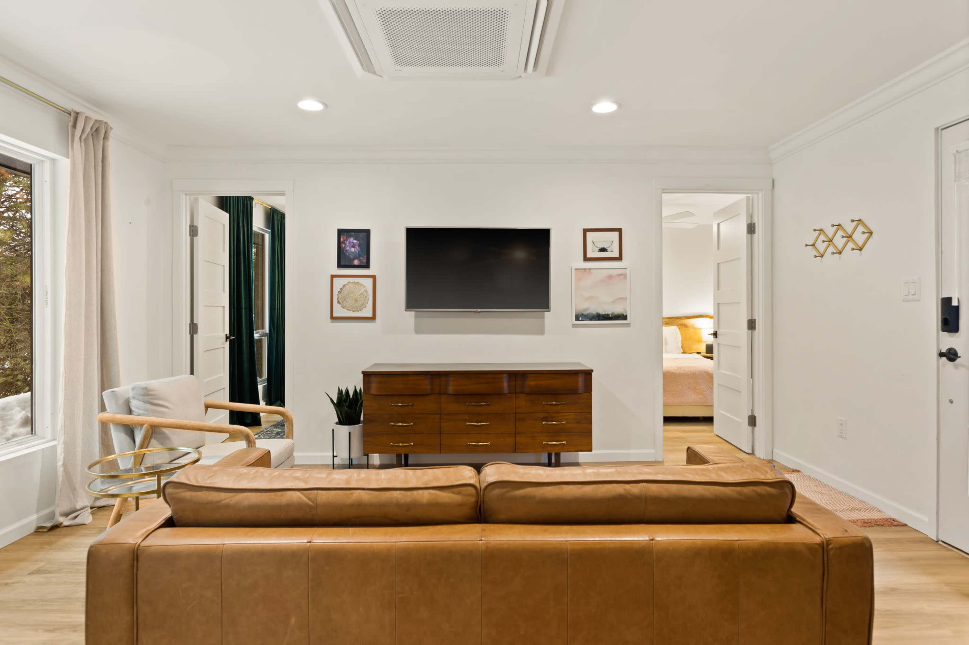 The image shows a living room with a brown leather sofa, a wooden TV stand, and a wall-mounted television, featuring a doorway leading to a bedroom in the background.