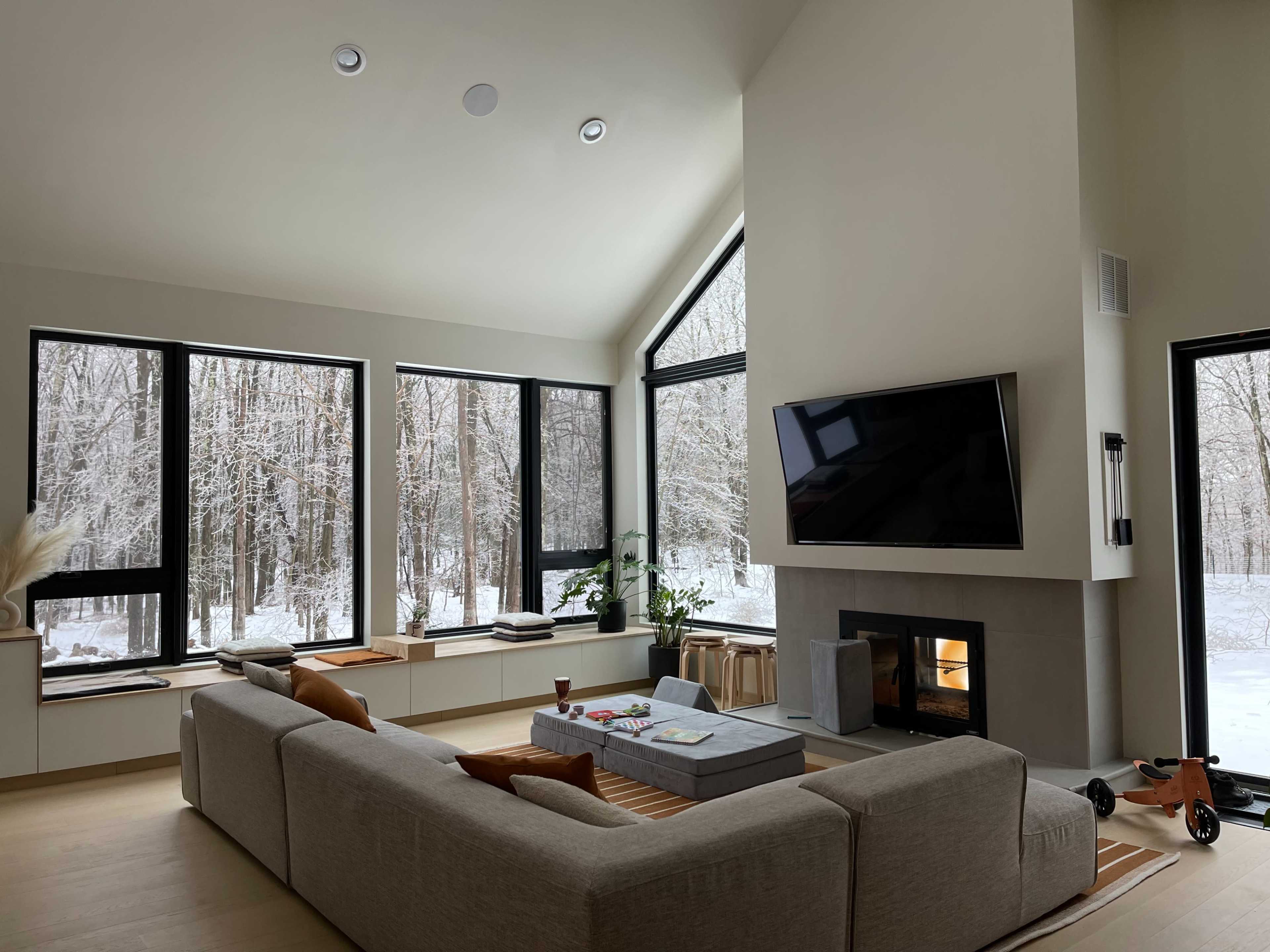 A modern living room features large windows overlooking a snowy forest, with a sectional sofa, a coffee table, and a wall-mounted television above a fireplace.