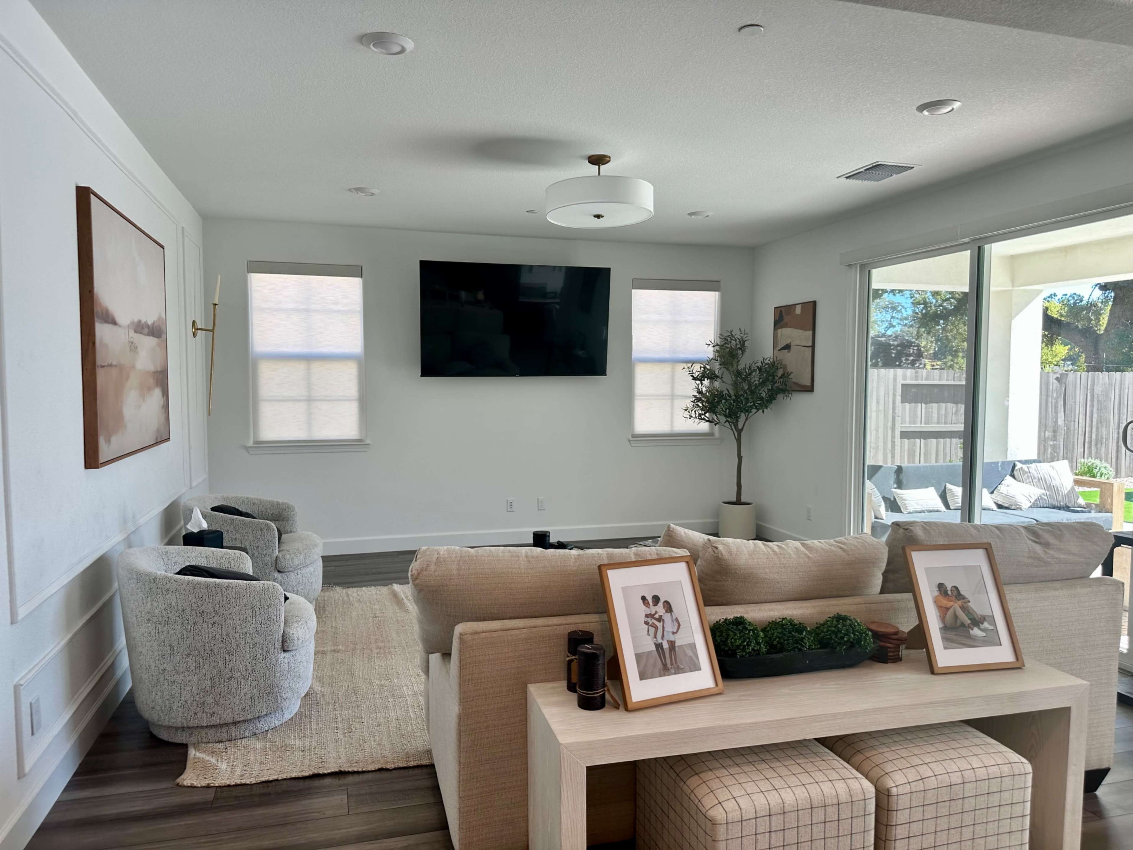 A modern living room features a neutral color palette, a large wall-mounted television, comfortable seating, and a sliding glass door leading to an outdoor area.