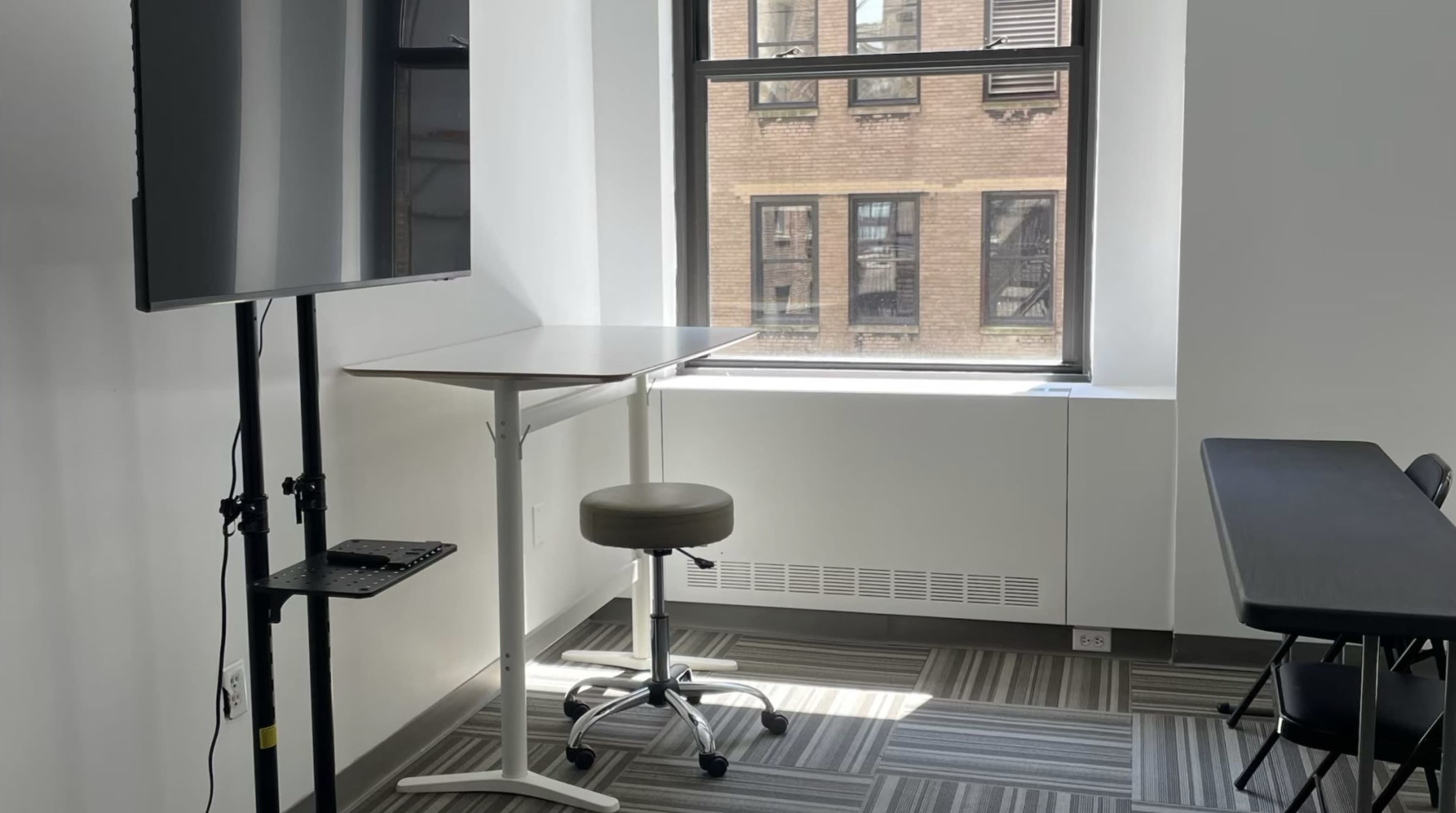 The image shows a small, modern office space featuring a high table with a stool, a wall-mounted screen, and a window overlooking a brick building.