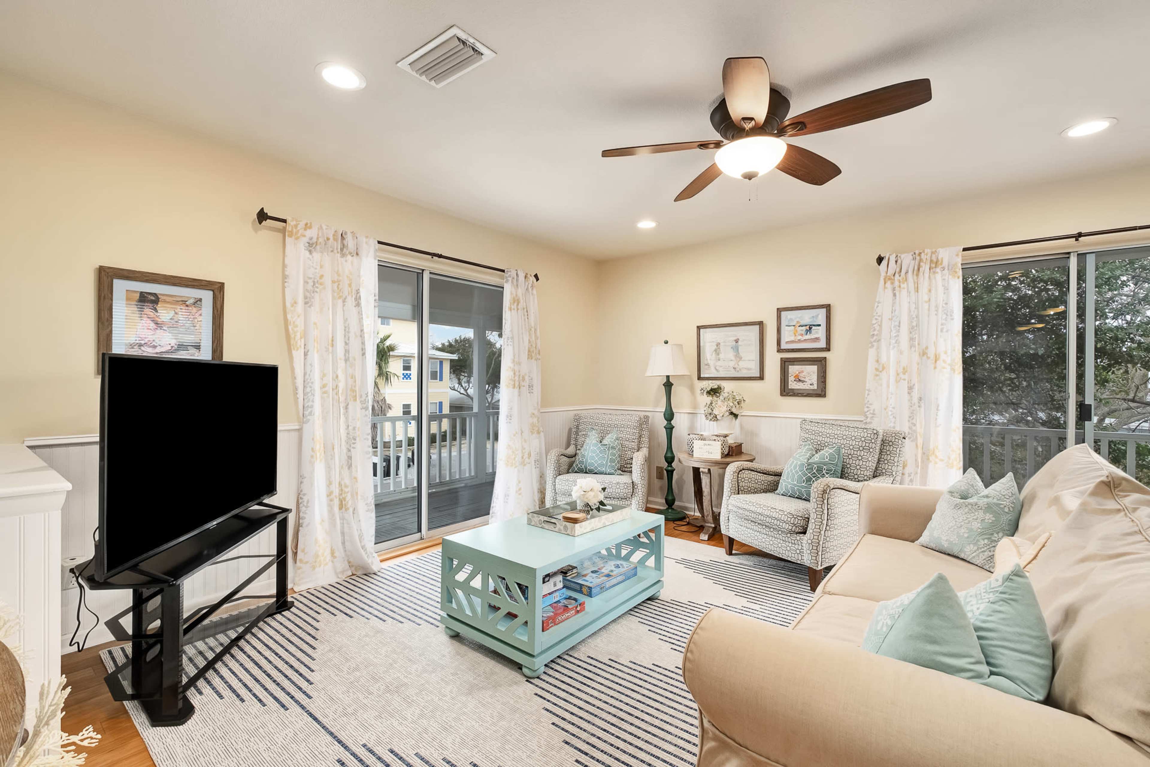A light-colored living room with a ceiling fan, a television on a stand, a coffee table, and comfortable seating arranged around large windows.
