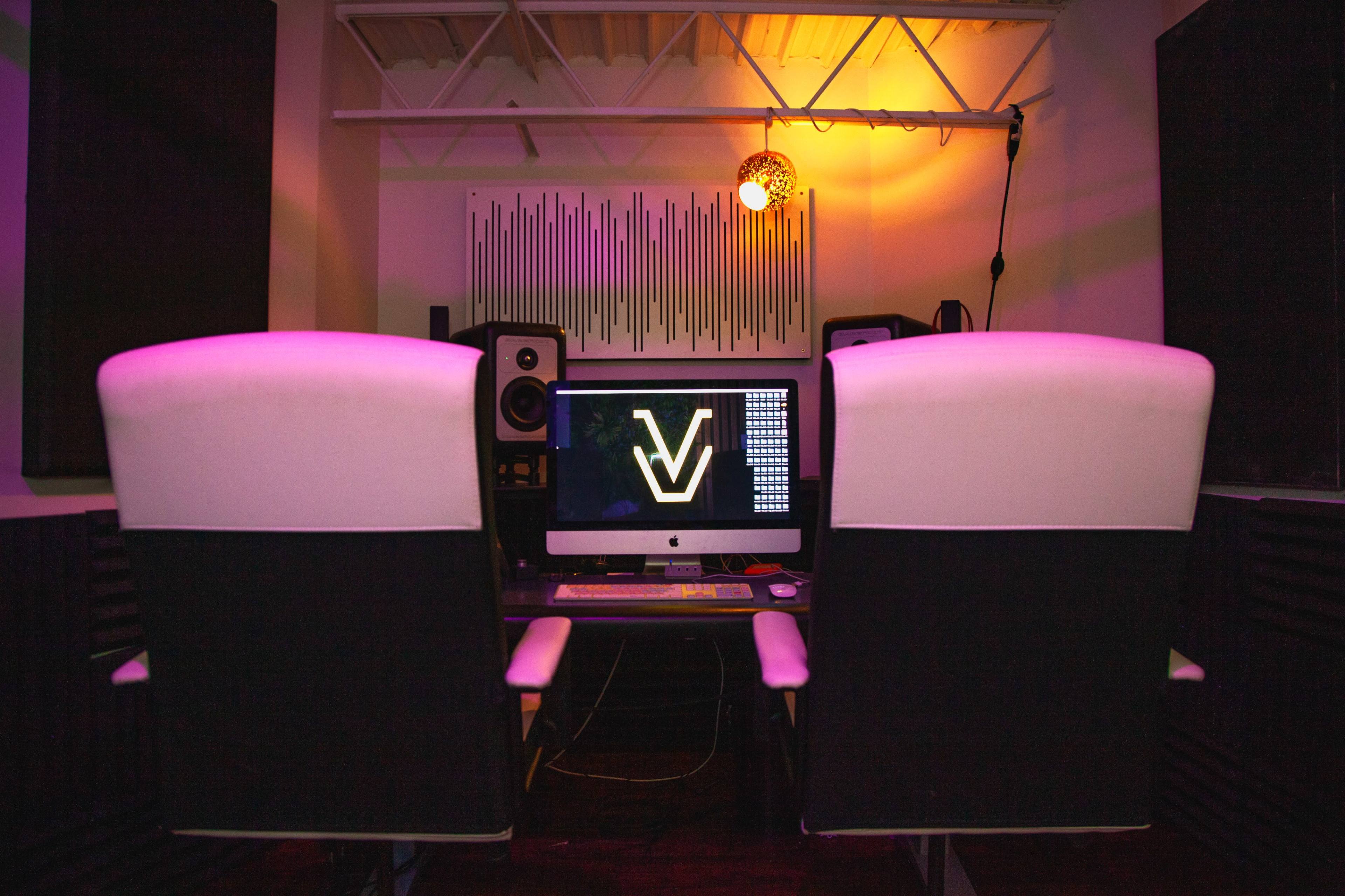 A music production studio with a computer desk featuring an Apple desktop and speakers, illuminated by colorful lighting.