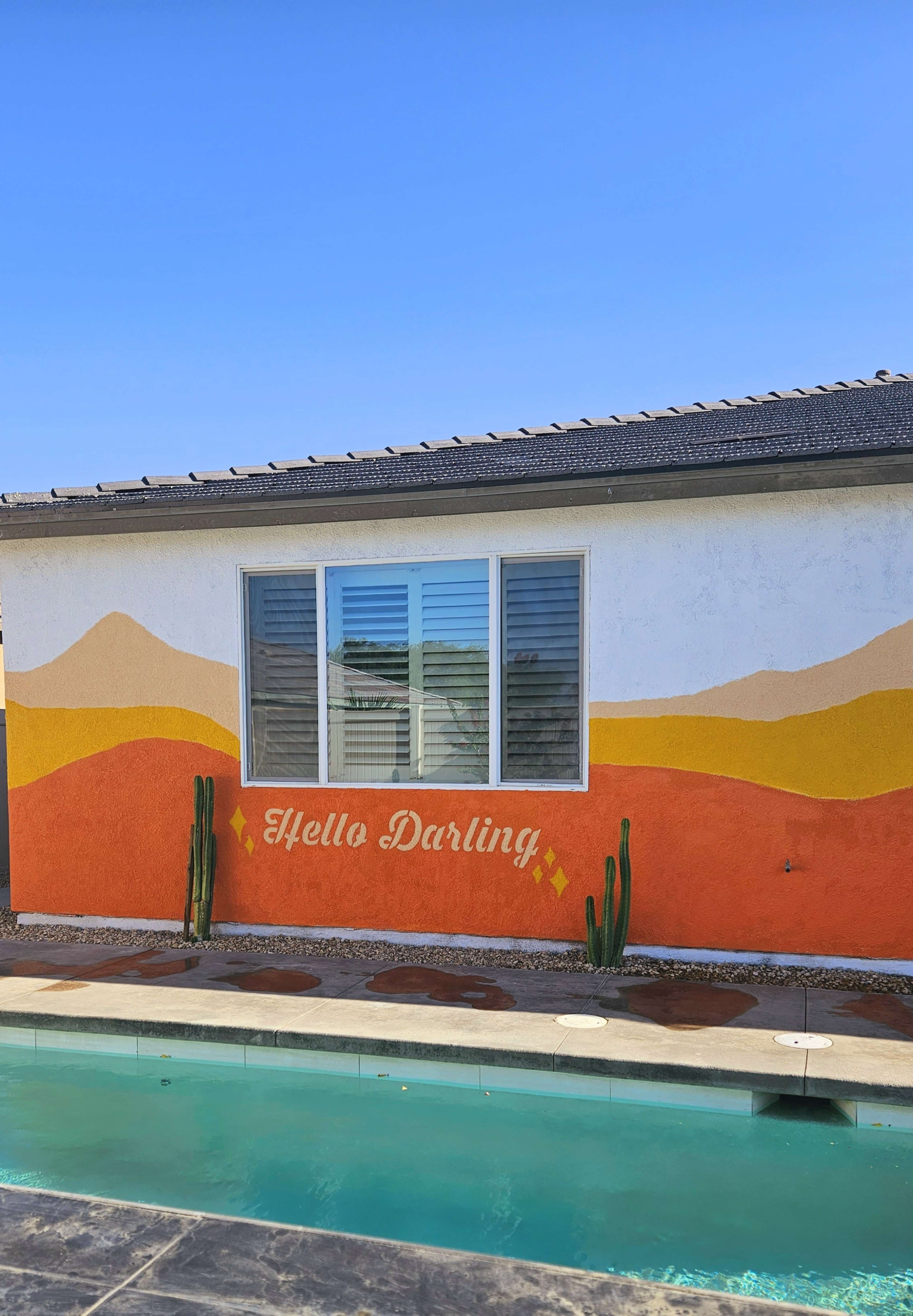 A brightly painted mural saying "Hello Darling" is featured on a white wall next to a swimming pool, with desert-themed designs and cacti in the foreground.