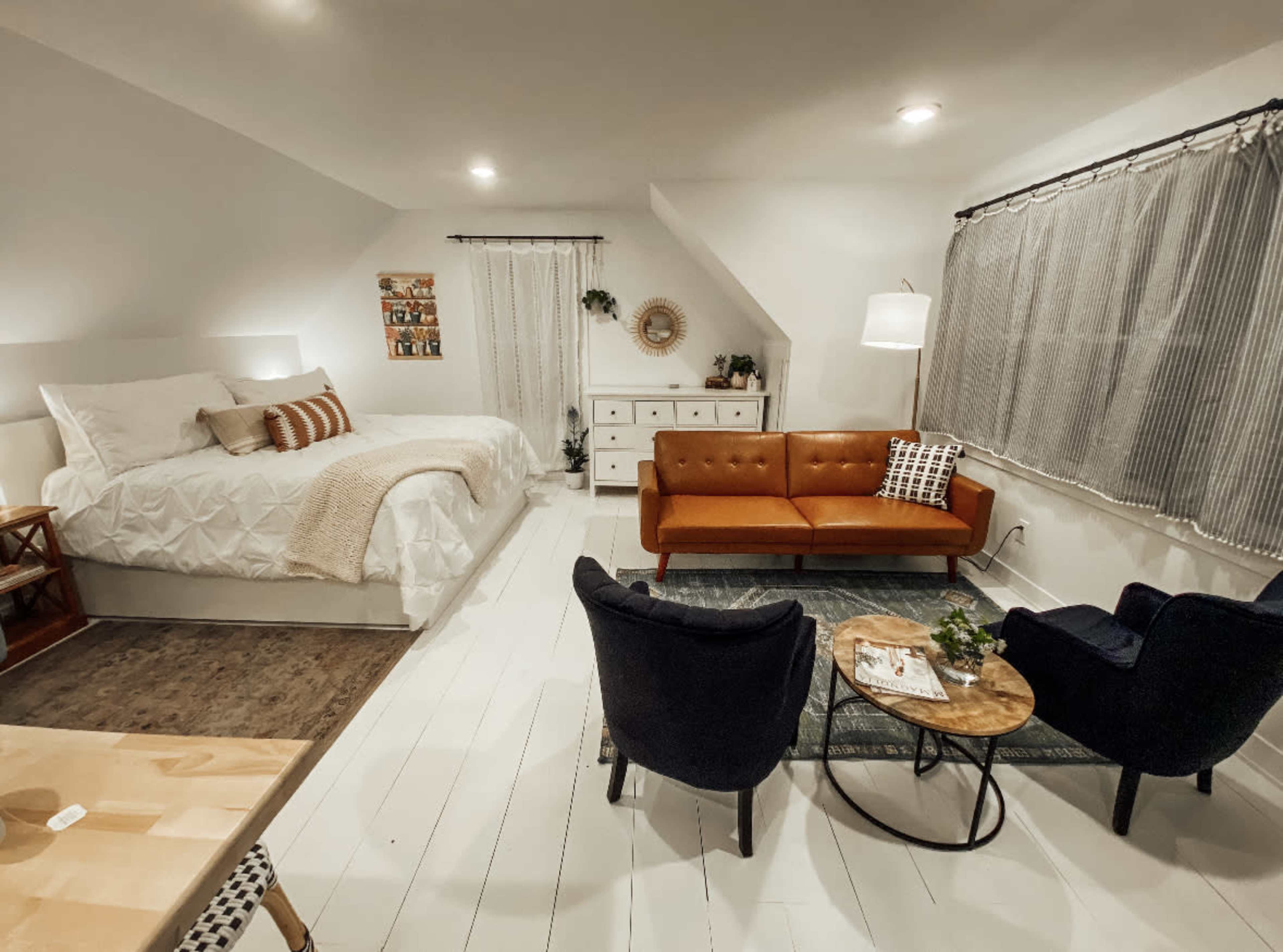 A cozy room divided into a sleeping area with a large bed and a living space featuring a brown sofa and two black armchairs around a circular table.