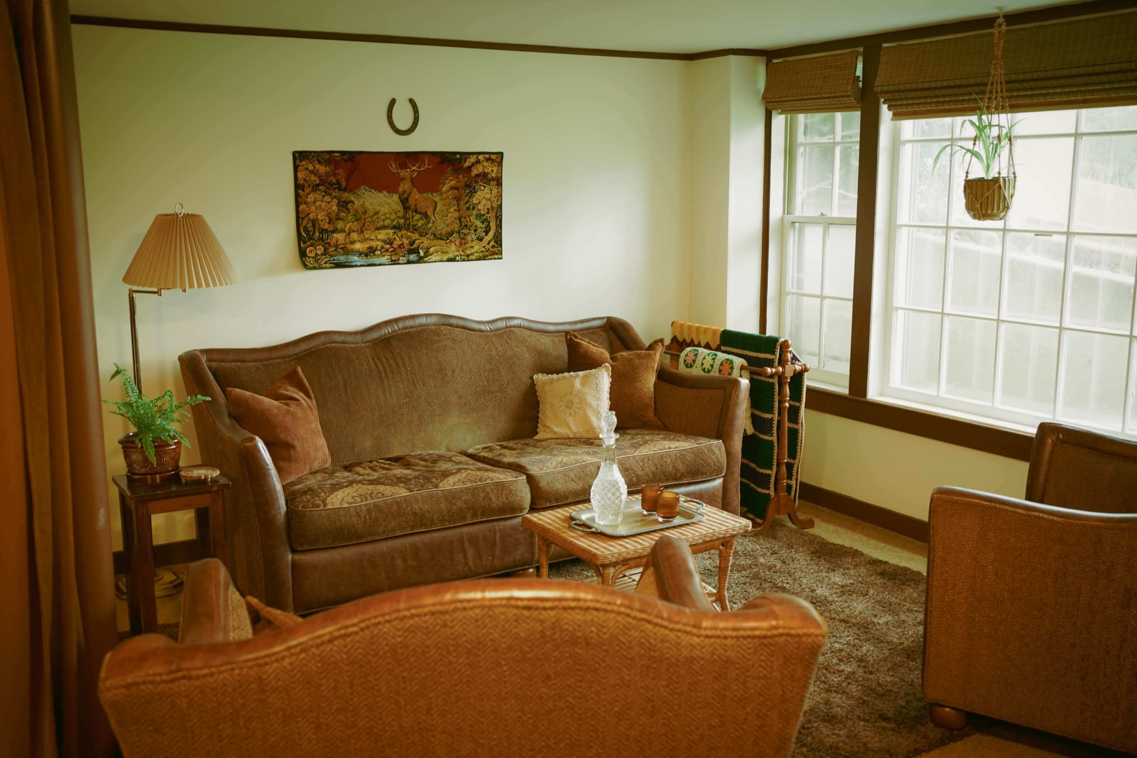 The image shows a cozy living room with a brown velvet sofa, two armchairs, a small wooden coffee table, and large windows allowing natural light in.