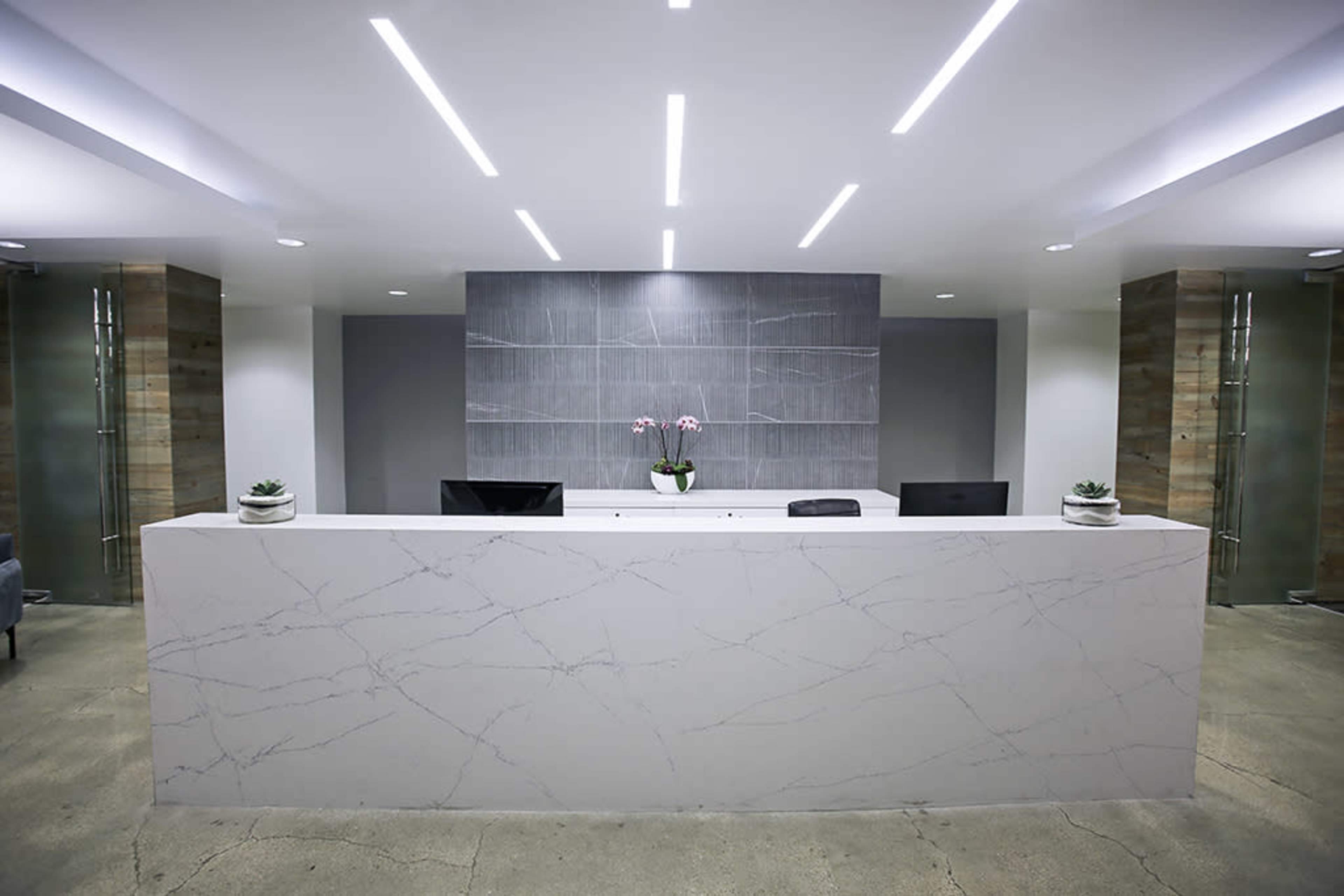 The image shows a modern reception area with a white marble desk, potted plants, and minimalist decor illuminated by sleek ceiling lights.