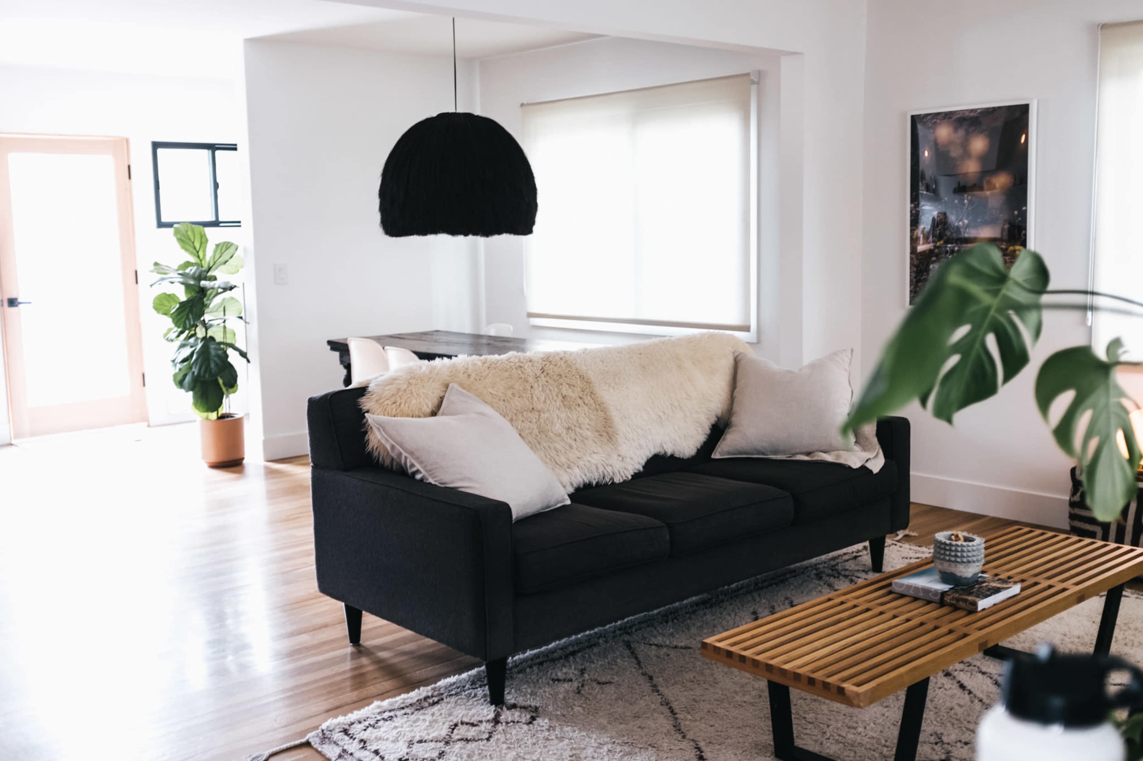 A modern living room features a dark sofa with a fluffy throw and pillows, accompanied by a wooden coffee table and potted plants.