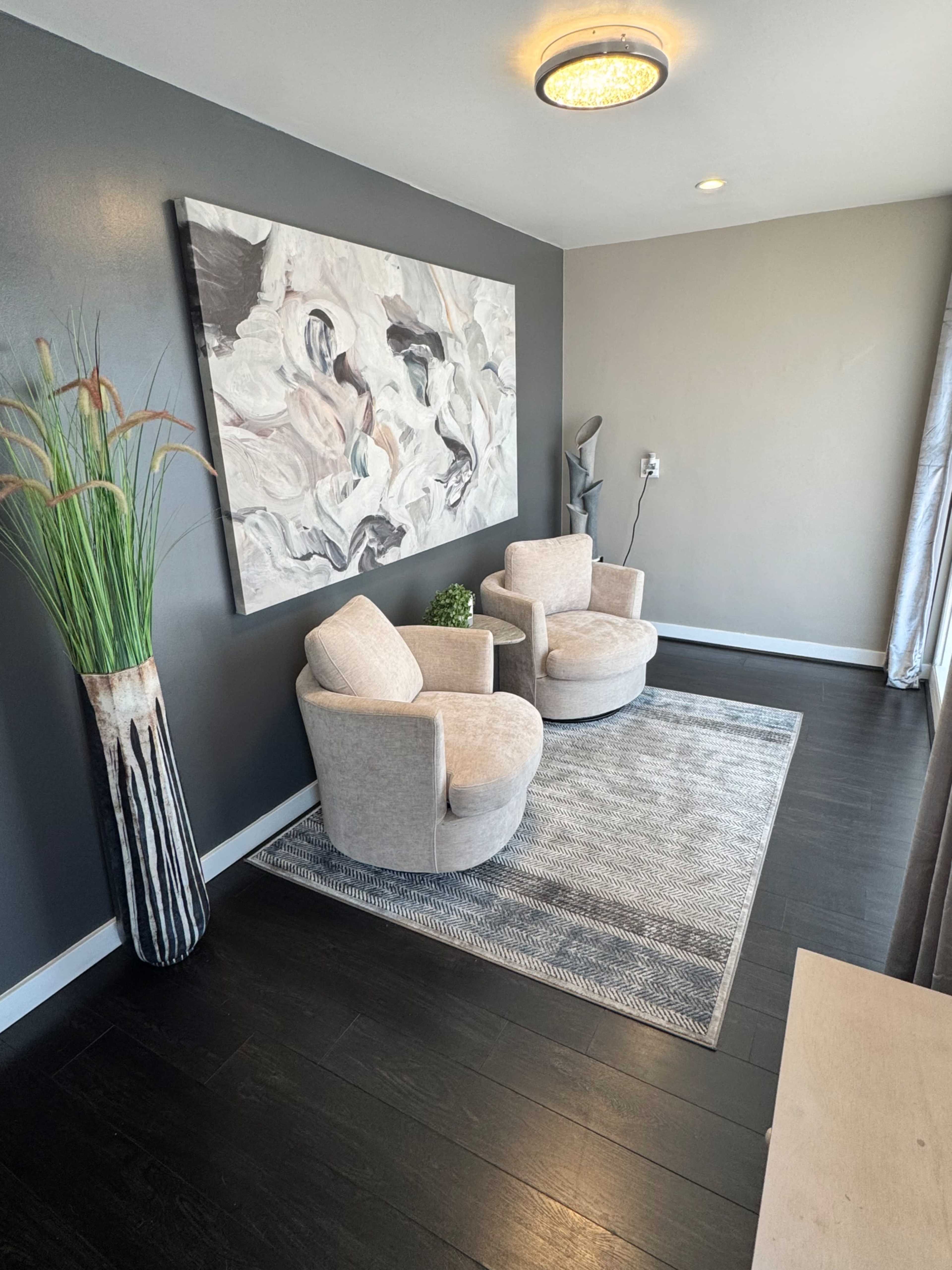 The image shows a modern interior space featuring two round, upholstered chairs beside a tall decorative plant and a large abstract painting on the wall.