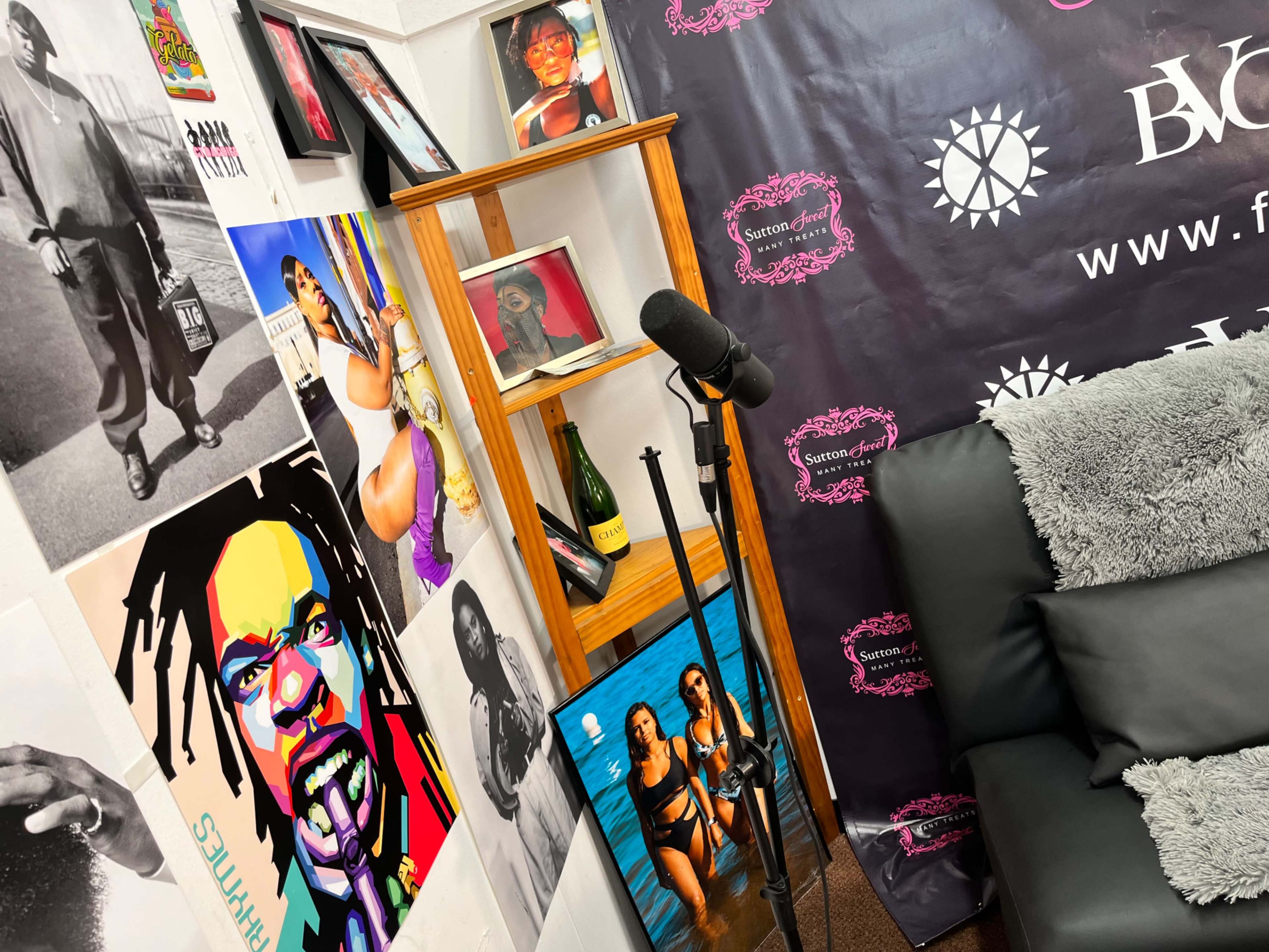A corner of a room with a wooden shelf displaying framed photos, a microphone on a stand, and decorative items, all against a backdrop of a patterned wall.