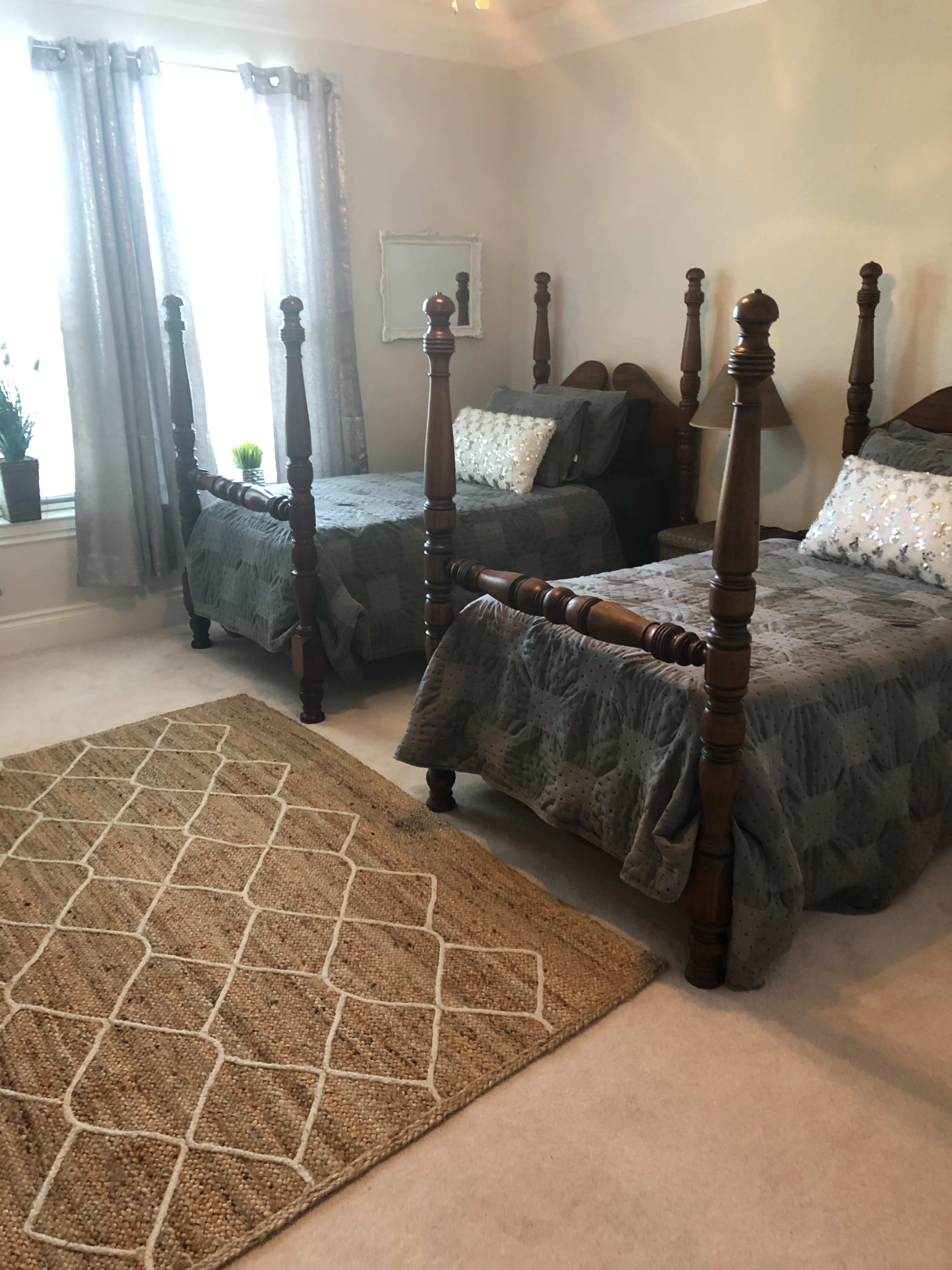 The image shows a bedroom with two wooden four-poster beds, a patterned area rug on the floor, and large windows with light curtains.