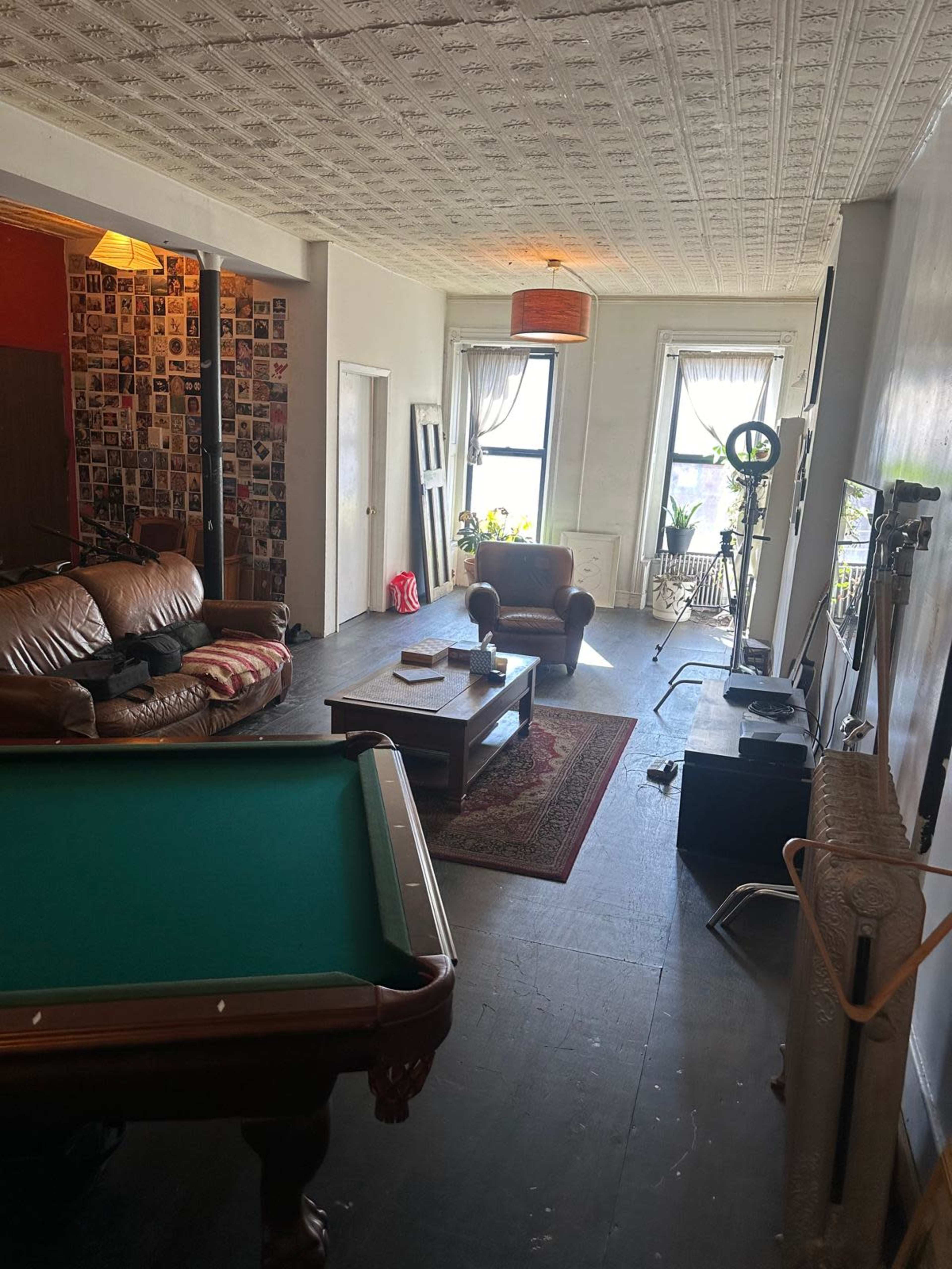 The image shows a spacious living room featuring a pool table, a brown leather sofa, a coffee table, and a television against the wall, with large windows allowing natural light.