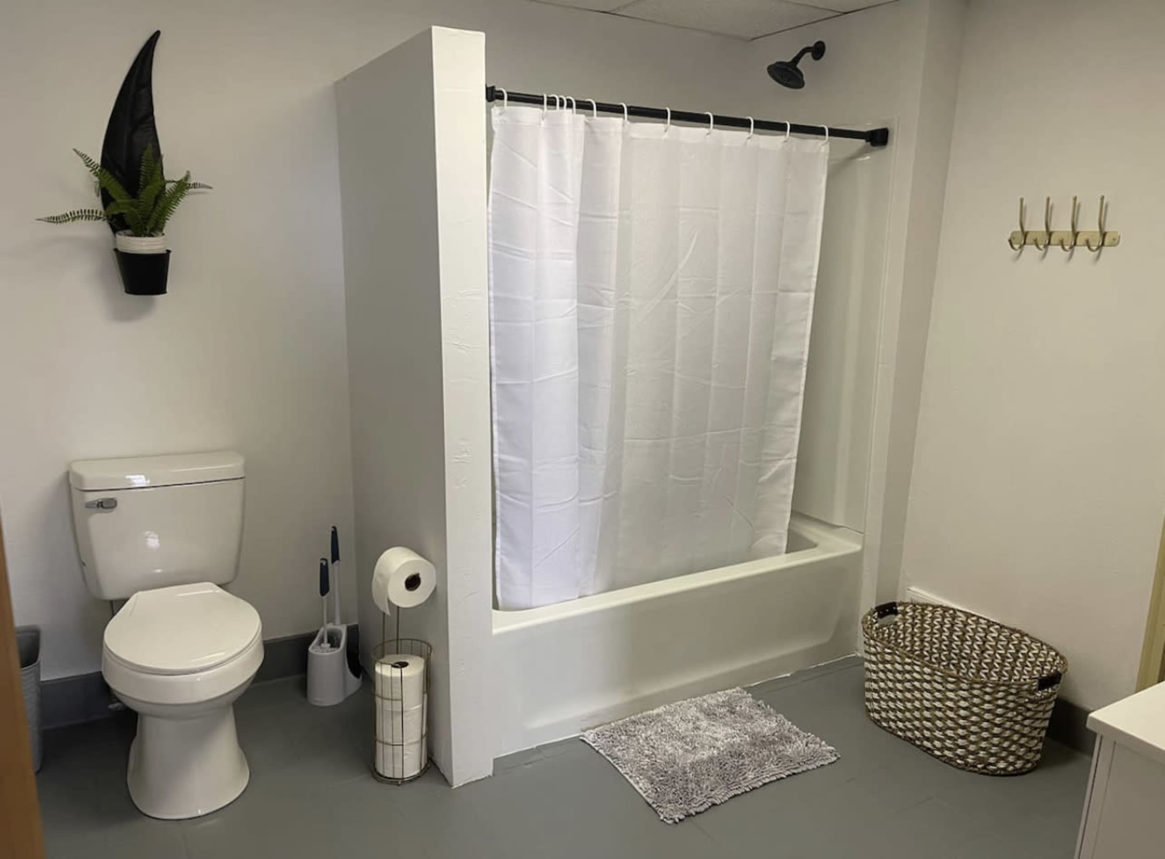 A bathroom featuring a toilet, a shower with a white curtain, a small mat on the floor, a waste basket, and a plant on the wall.