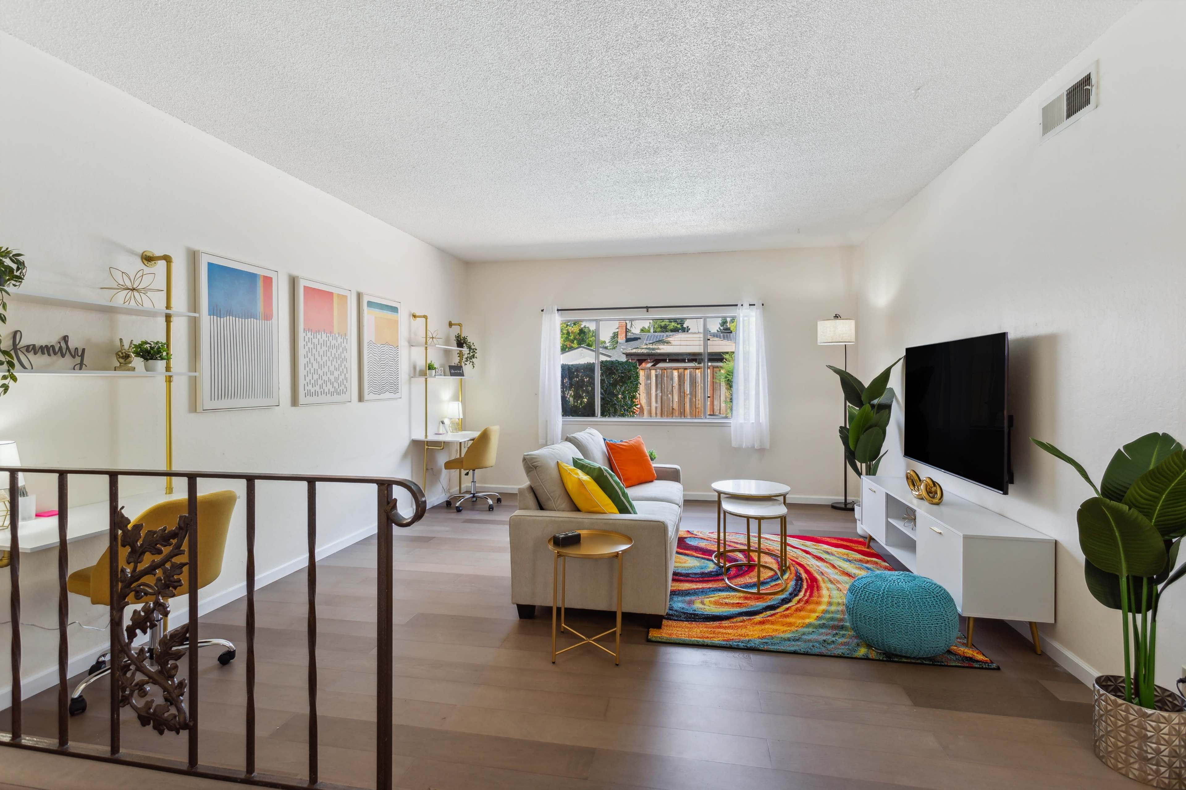 A brightly lit living room featuring a sofa, a coffee table, a colorful rug, and a television, with a workspace and large window overlooking a patio area.