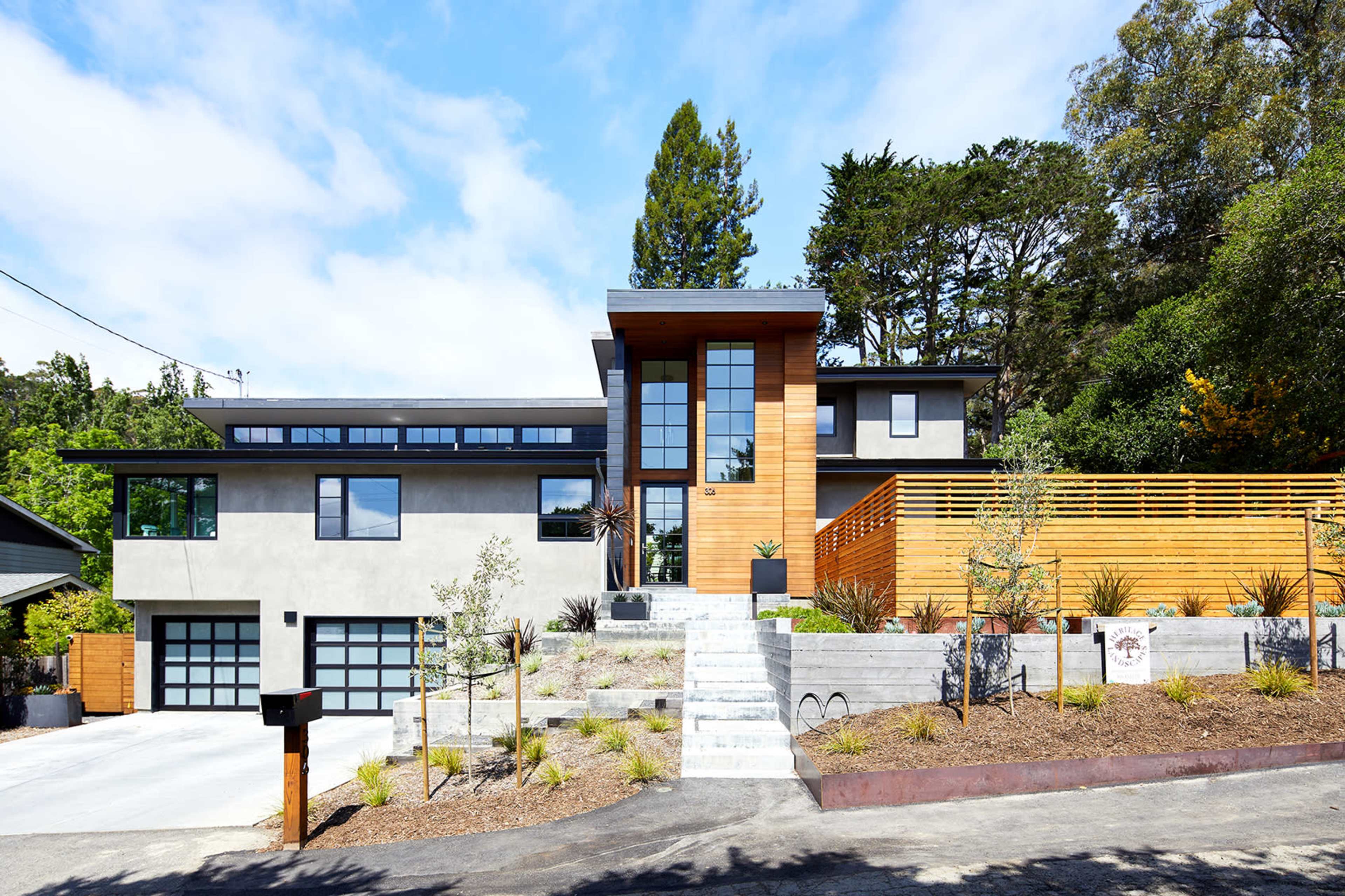 The image shows a modern two-story house with a wooden entrance, large windows, and a landscaped front yard featuring steps and planters.