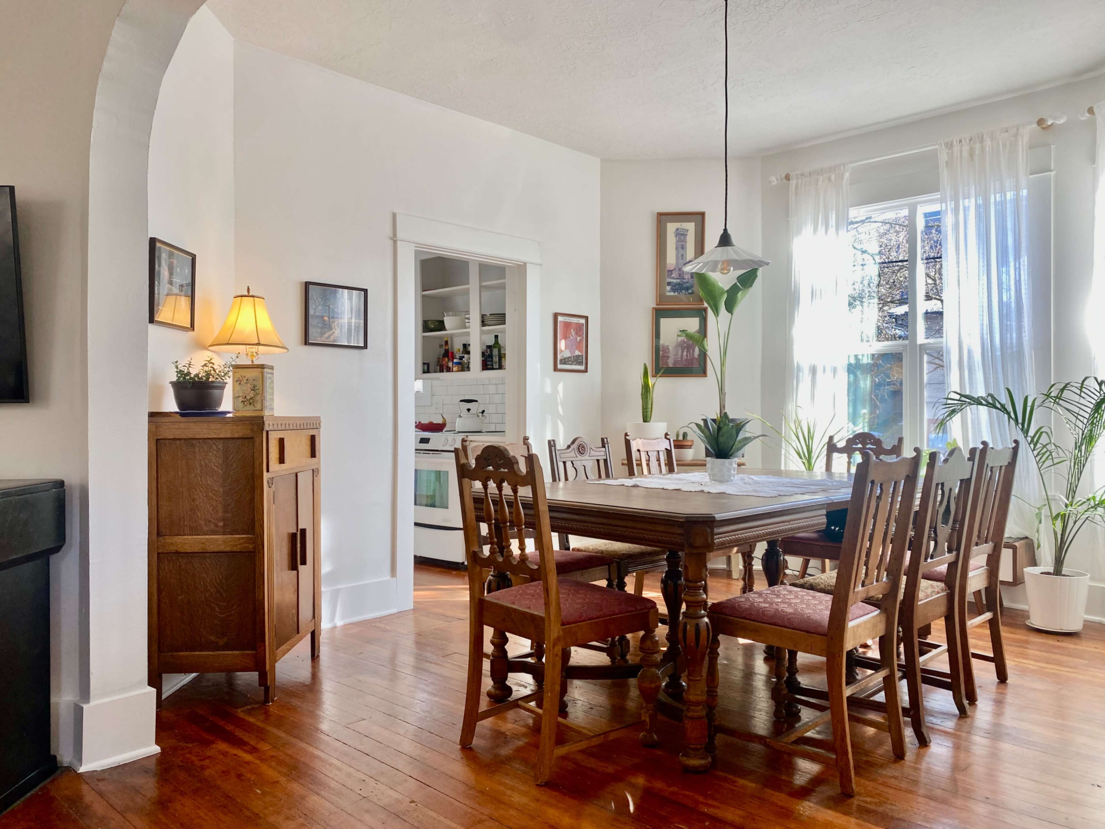 A wooden dining table with chairs is set in a bright room featuring large windows and plants, with a kitchen visible in the background.