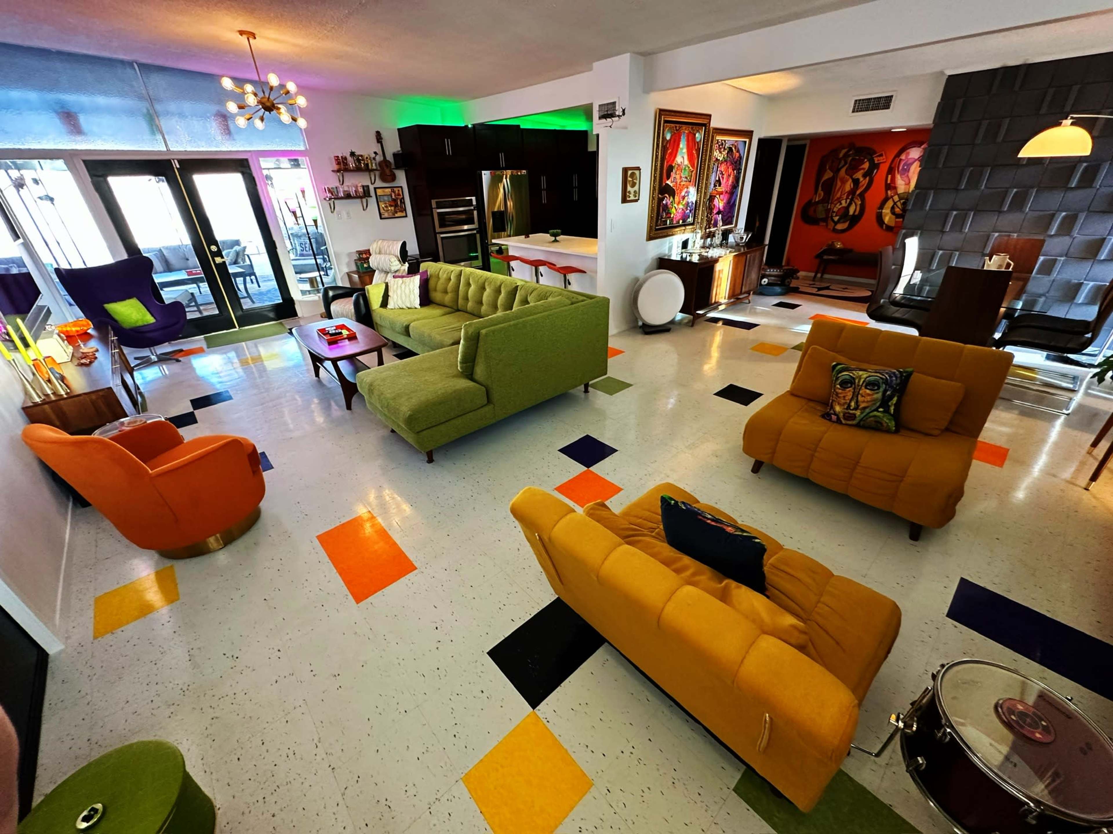 The image shows a vibrant living area with colorful furniture, including green, orange, and yellow sofas, a round coffee table, and artwork on the walls.