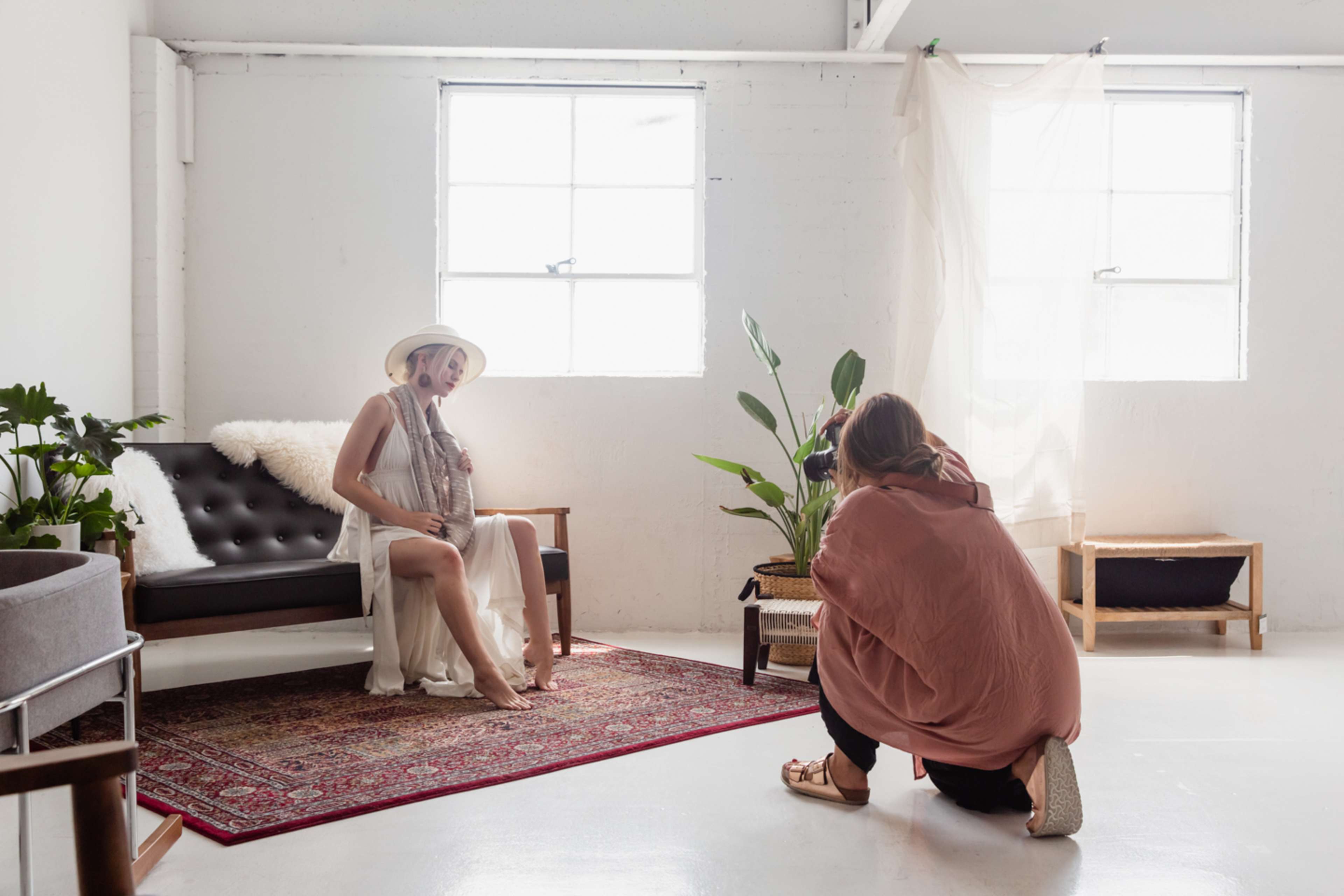 A photographer captures a model sitting on a couch in a bright, minimalist studio with large windows and indoor plants.