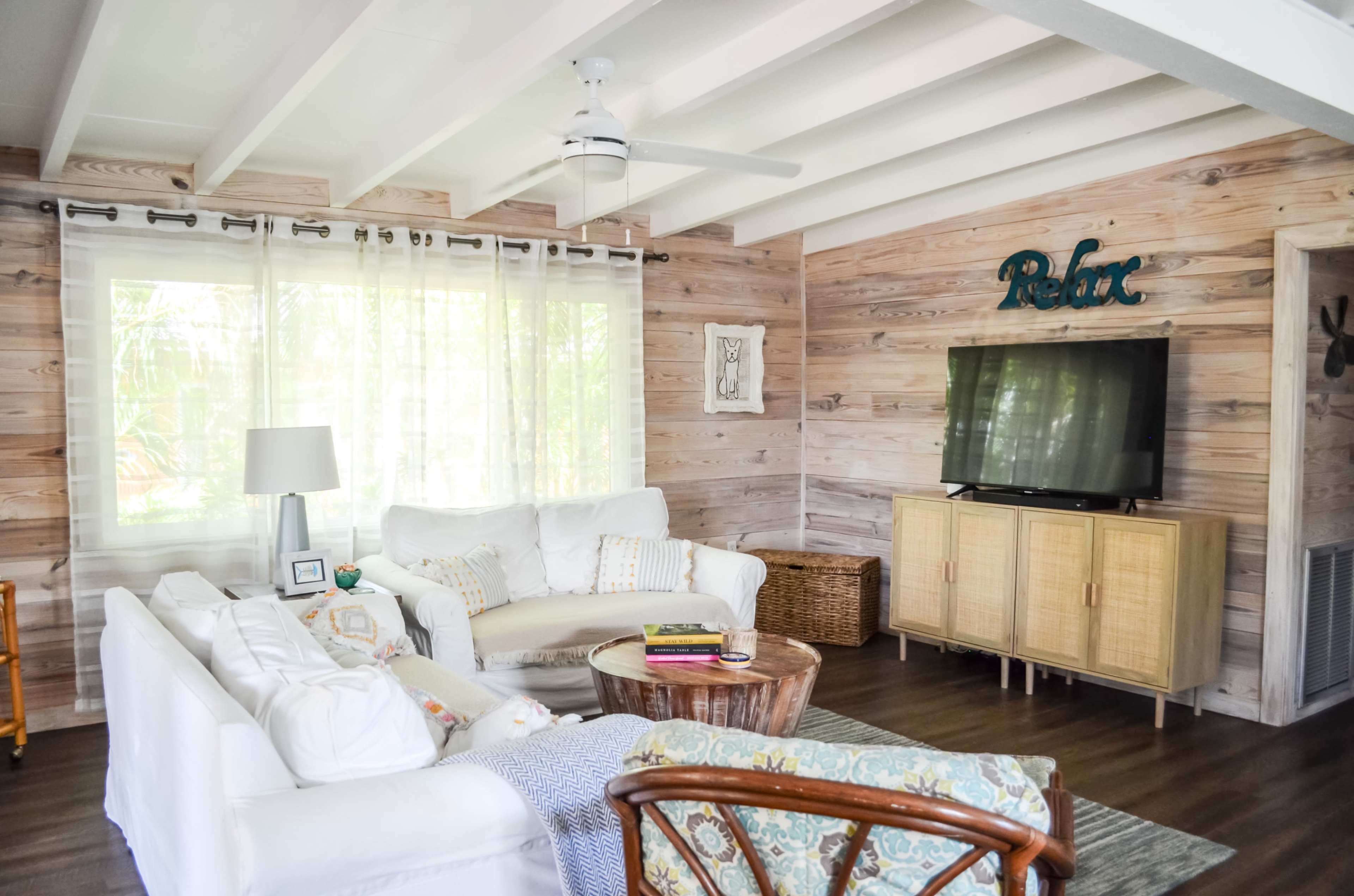 The living room features white sofas, a wooden coffee table, a TV on a cabinet, and a large window with sheer curtains, all set against a backdrop of wooden paneling.