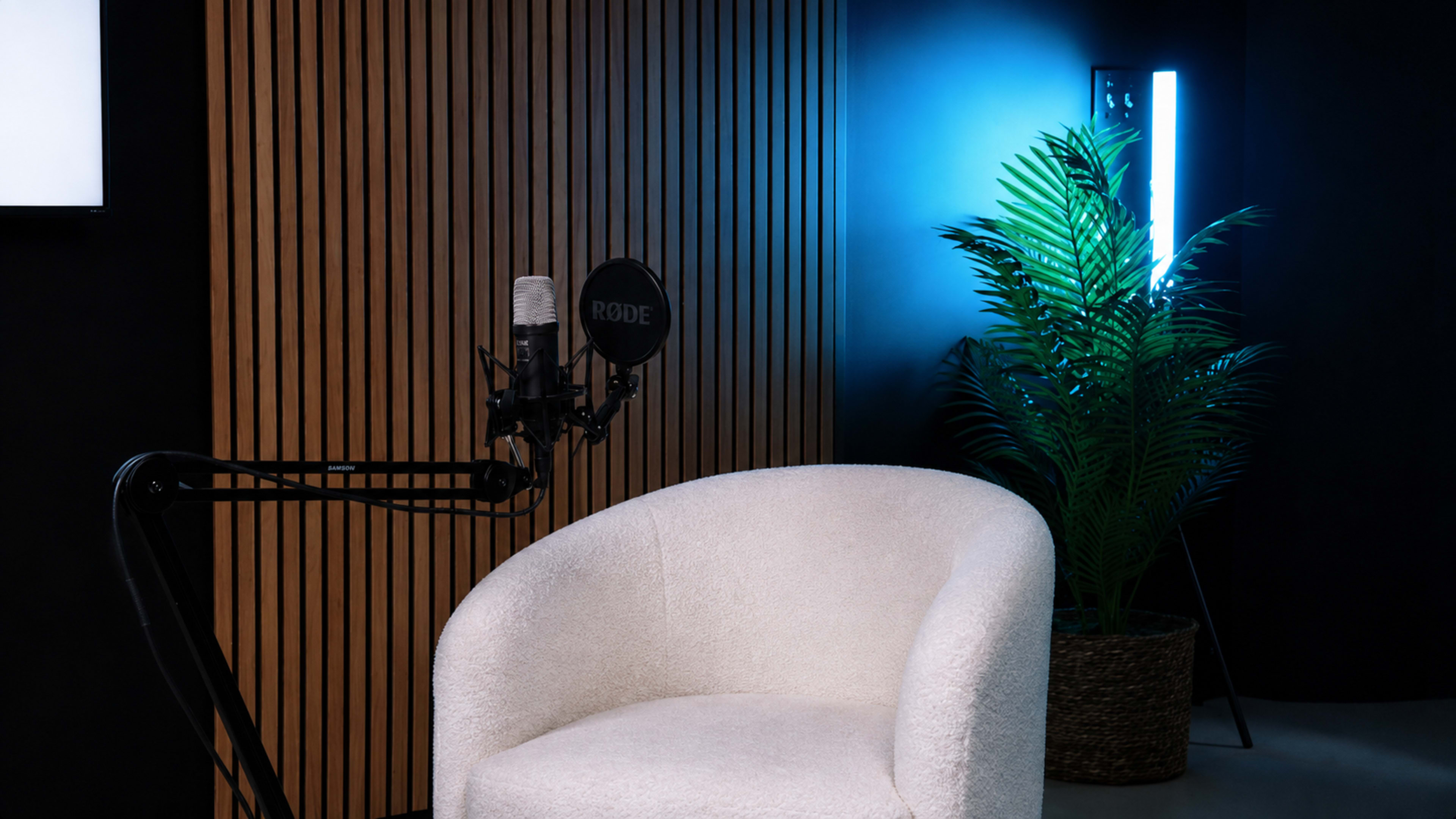 The image shows a cozy studio setup featuring a microphone mounted on an arm, a white chair, a potted plant, and a contrasting illuminated backdrop.