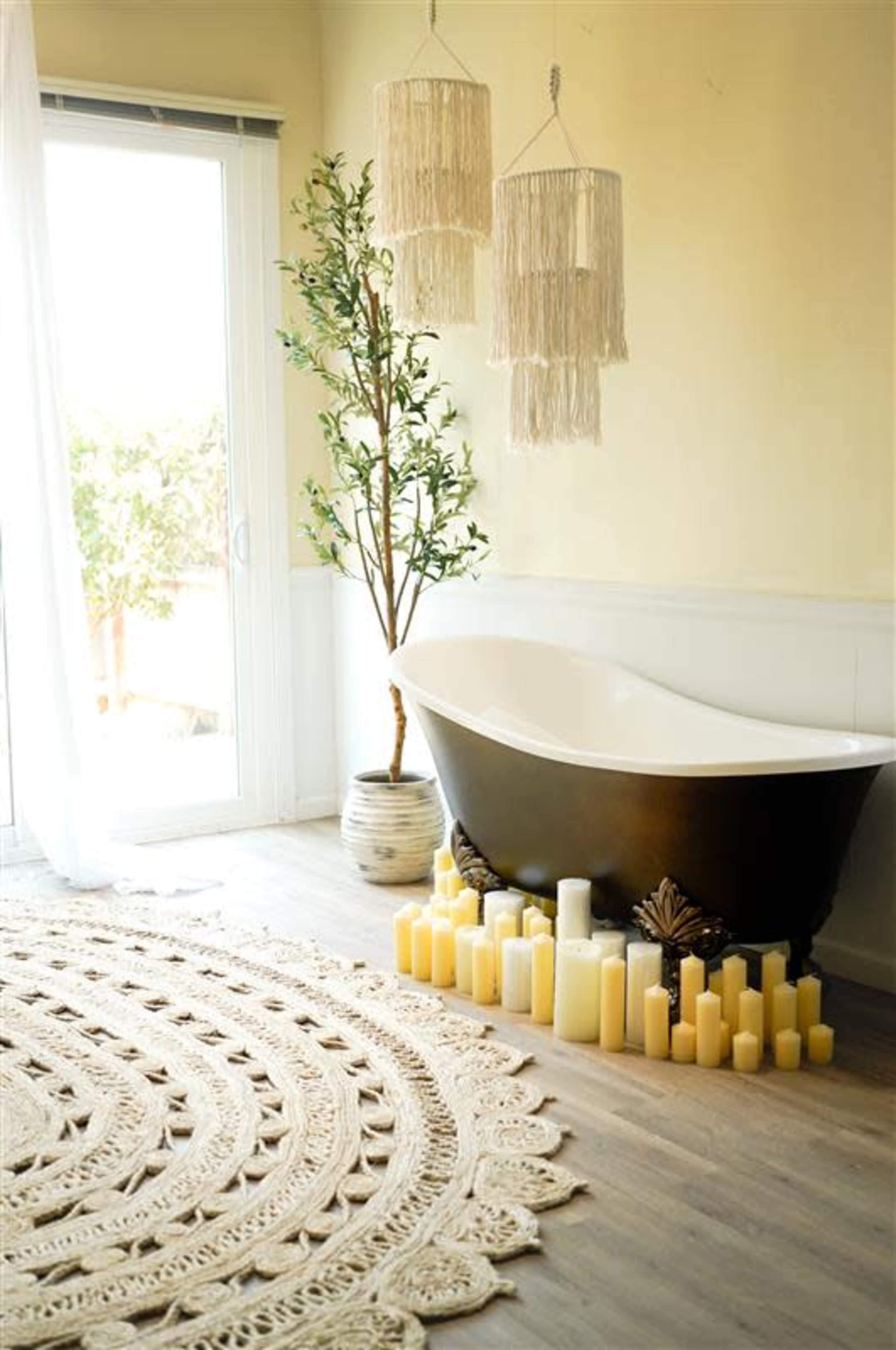 A freestanding black bathtub is positioned beside a large window, surrounded by a circular decorative rug and multiple cream-colored candles.