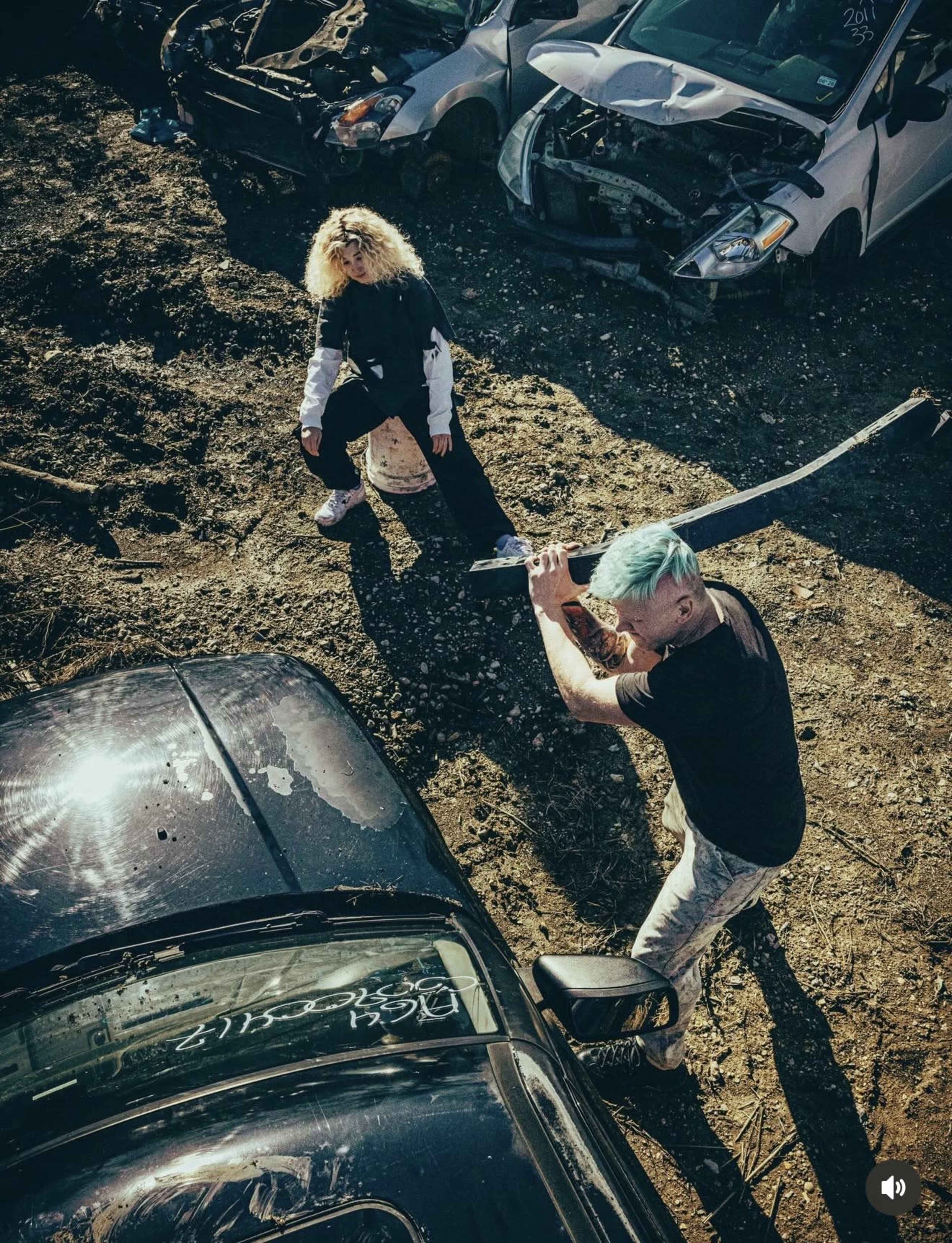 A person with blue hair raises a weapon while another person stands nearby amidst wrecked cars in a junkyard.