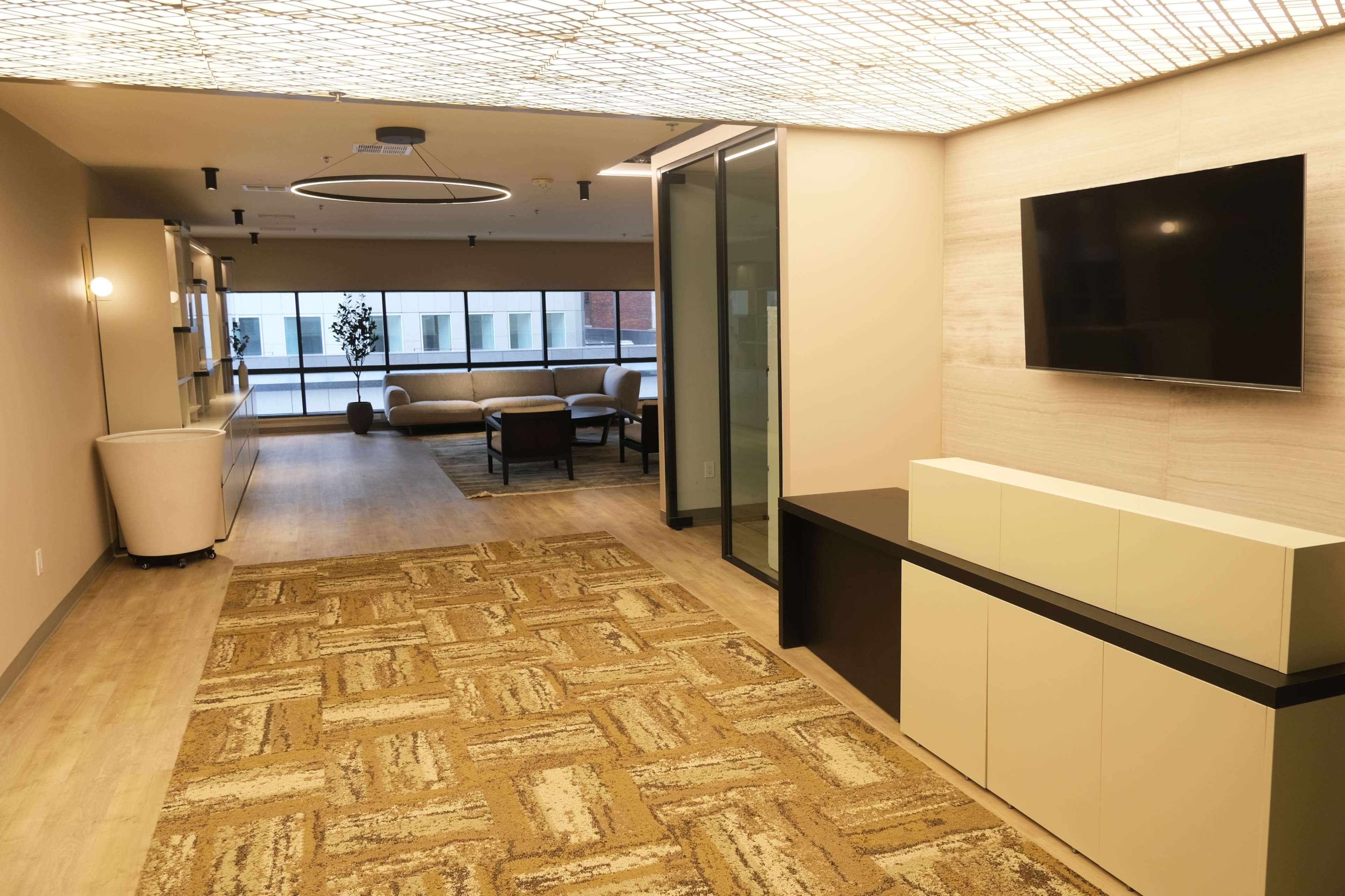 A modern office space with a carpeted area, a television mounted on a wall, and a seating arrangement visible through a glass partition.