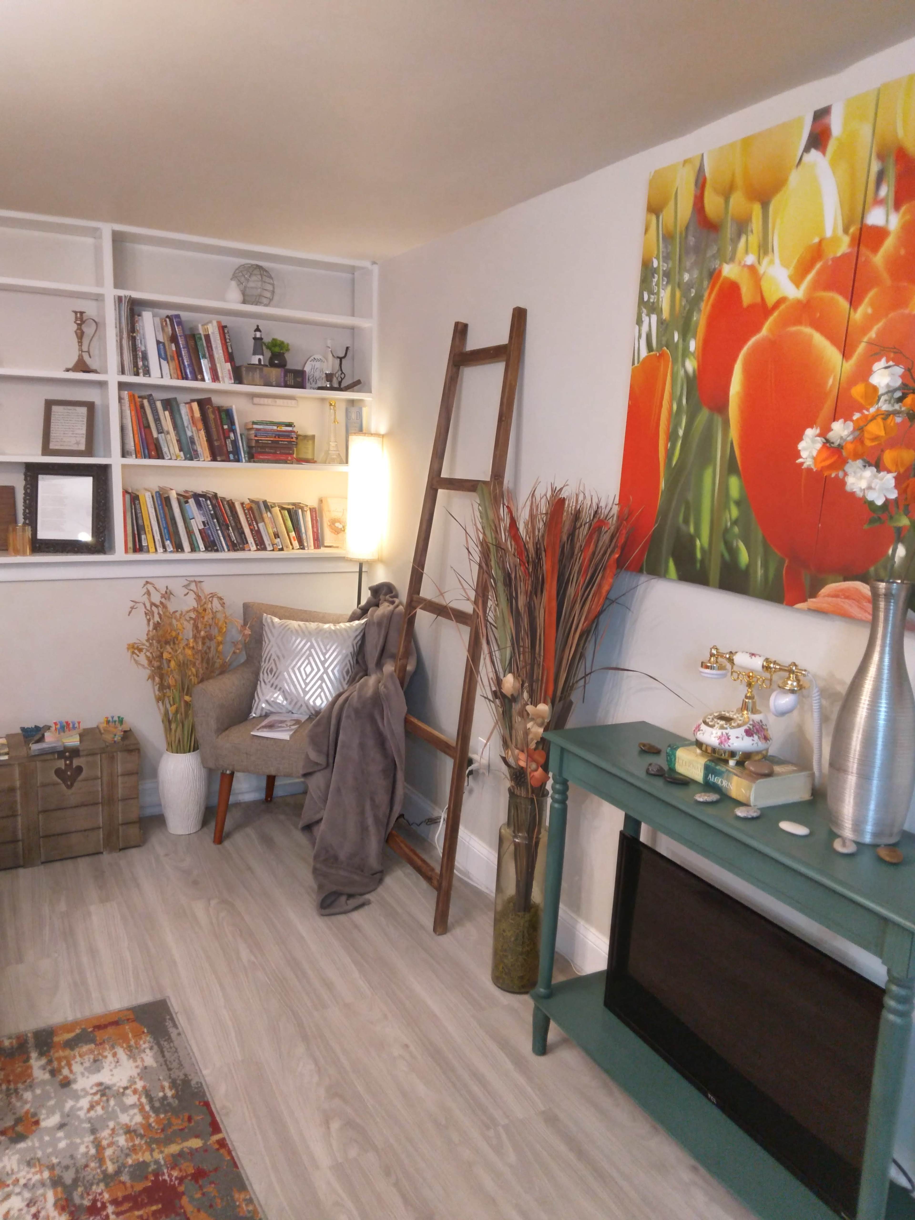 A cozy living area featuring a bookshelf, a decorative ladder, a green console table, ornamental plants, and a vibrant flower painting on the wall.