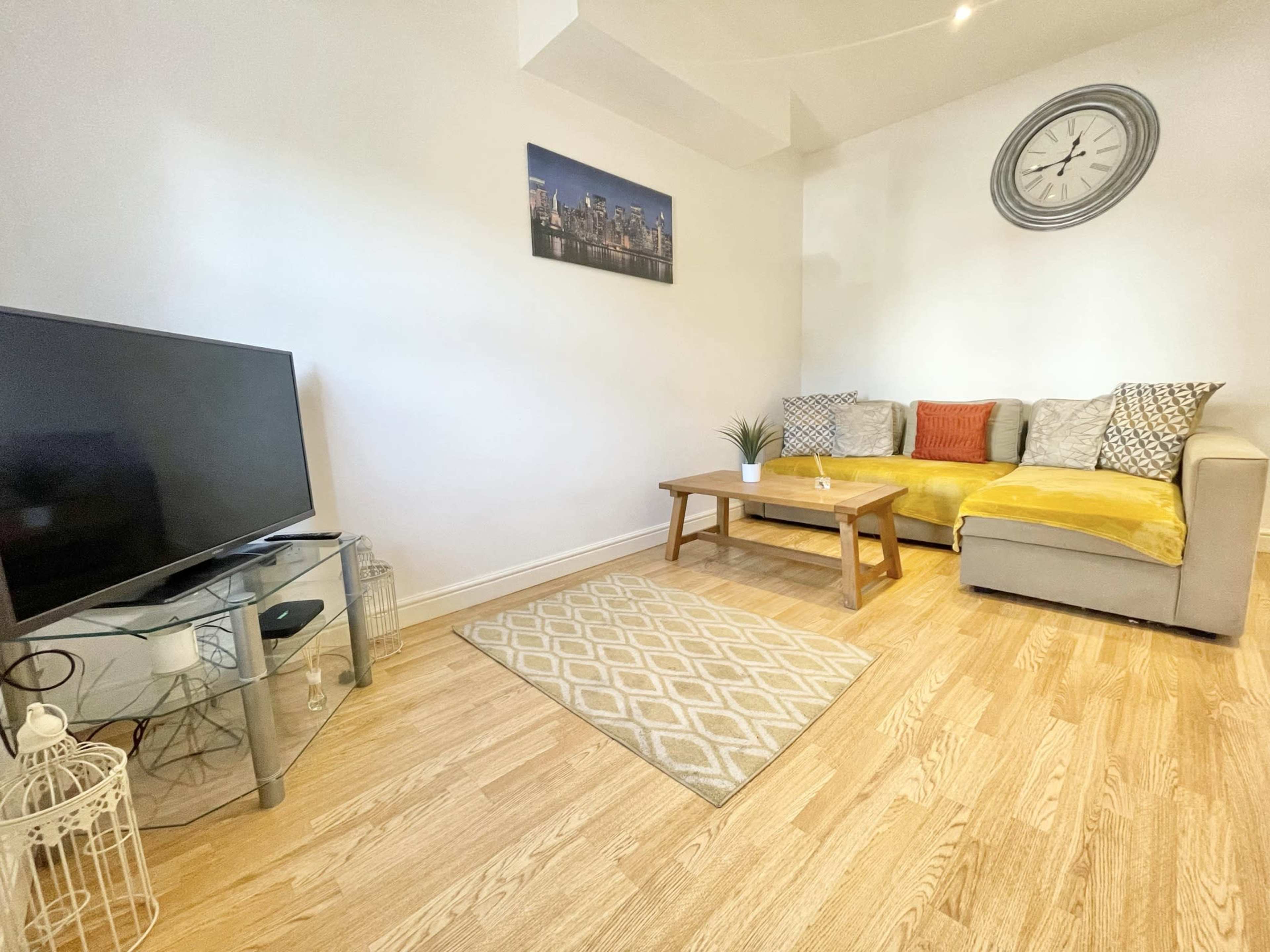 A living room with a corner sofa, a coffee table on a patterned rug, a television on a glass stand, and a wall clock above.