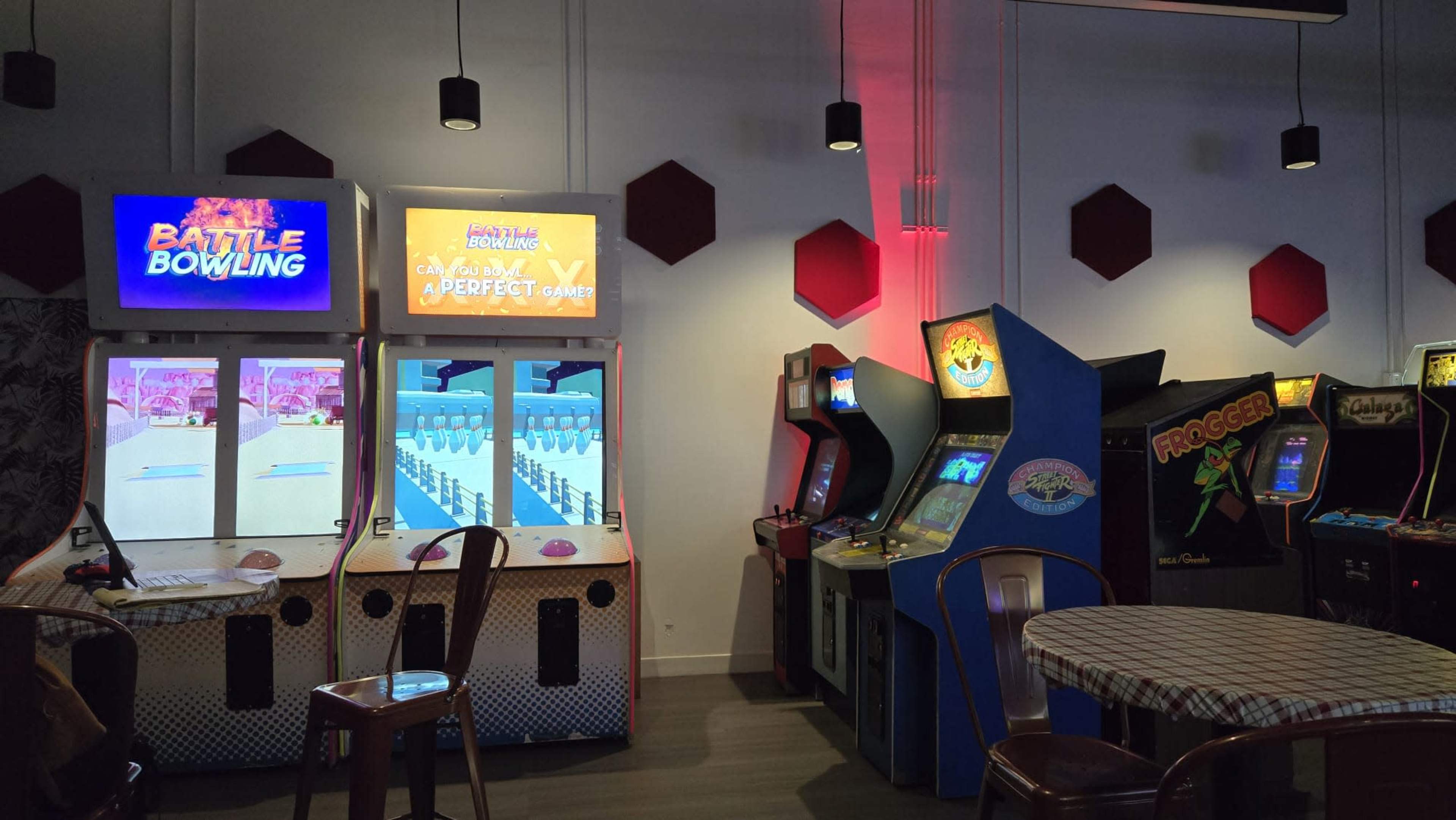 The image shows a retro arcade with several gaming machines, including "Battle Bowling" and "Froggin'," set against a backdrop of red hexagonal wall decorations.