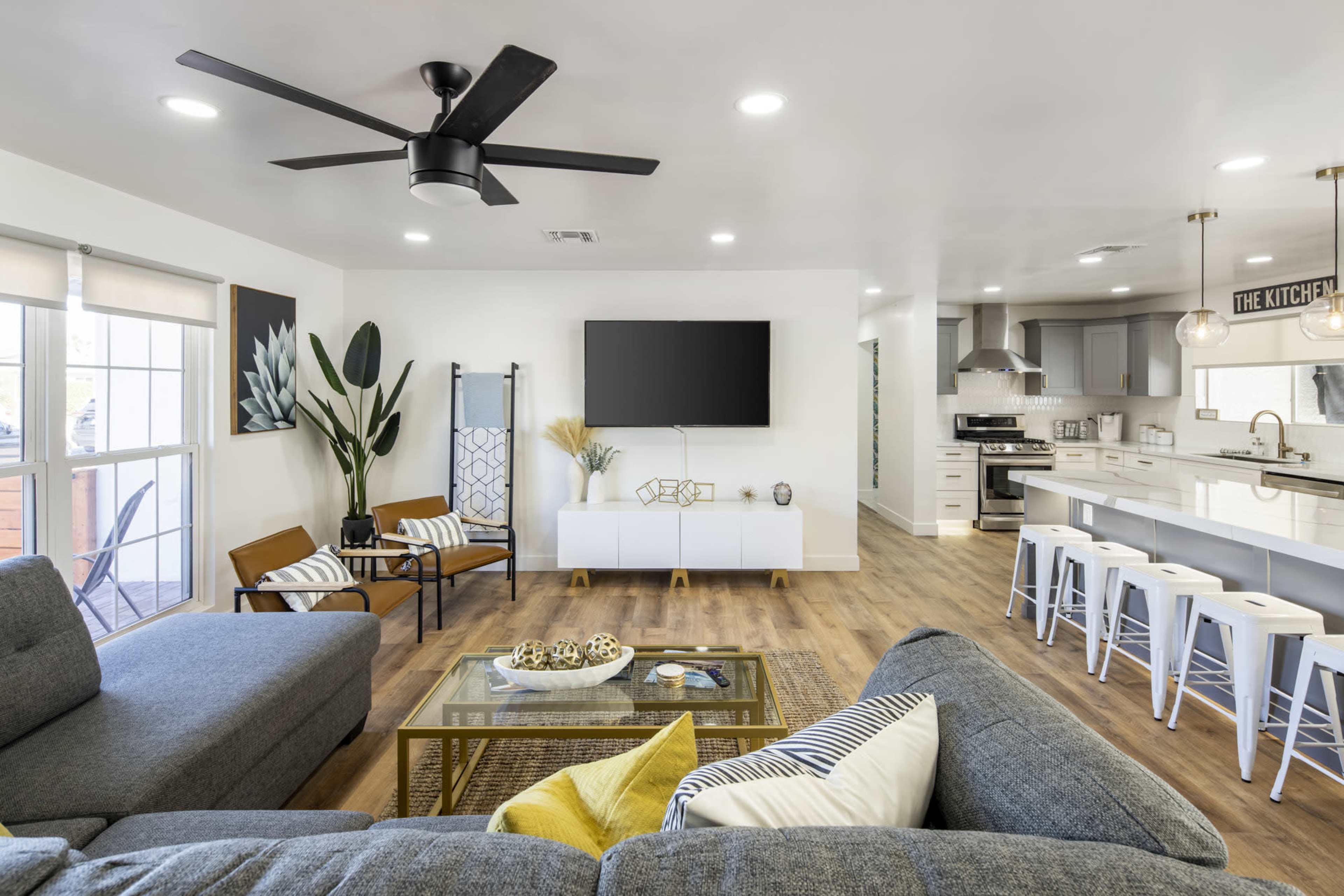 A modern living room with a gray sectional sofa, a large wall-mounted television, and an open kitchen area featuring white cabinetry and bar stools.