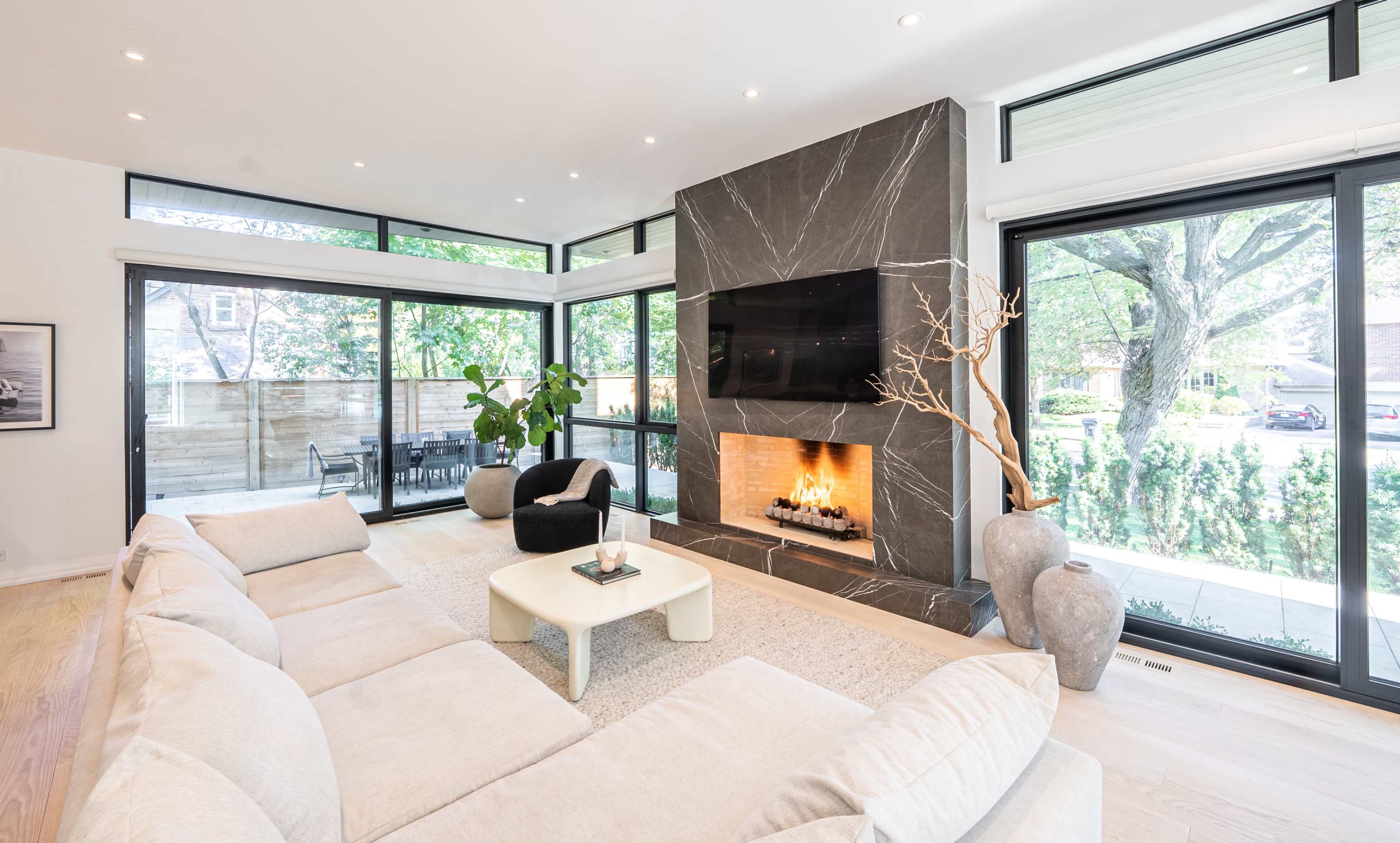 A spacious living room features large windows, a modern fireplace with a TV above it, and a light-colored sectional sofa.