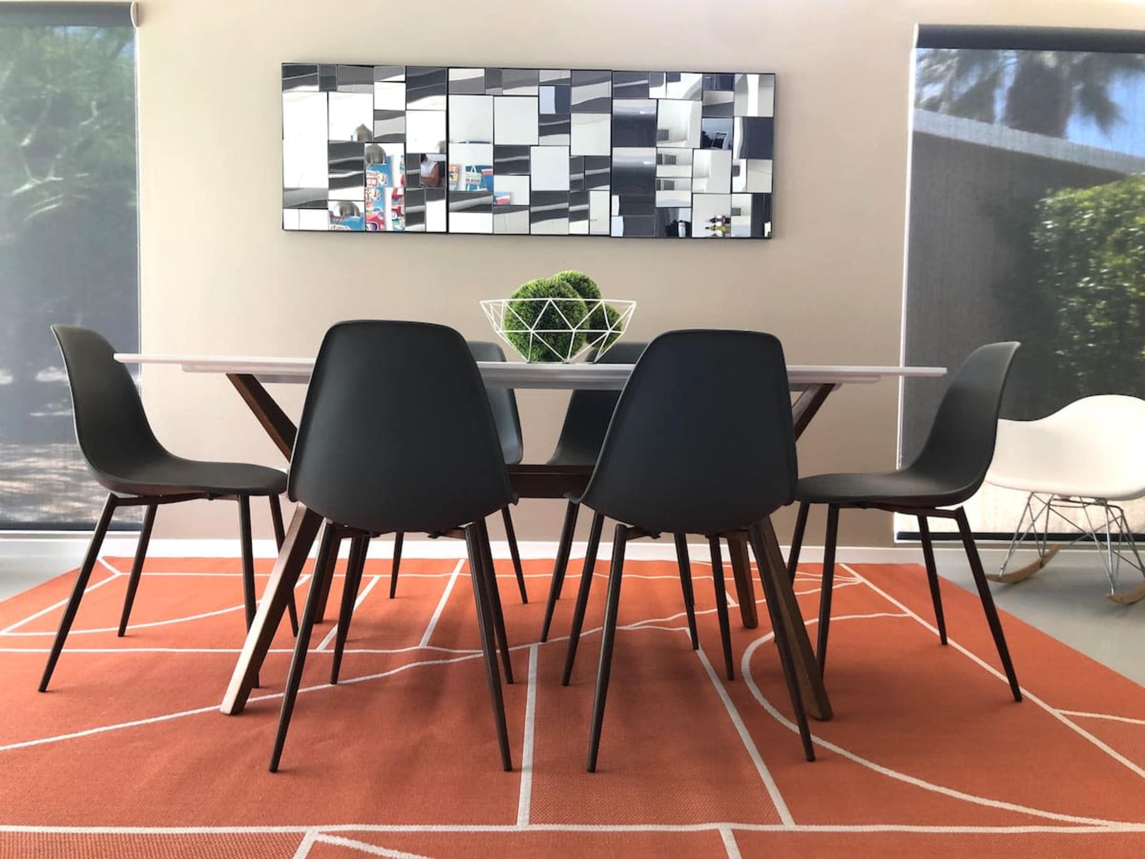 A dining table with a white top is surrounded by five black chairs on an orange rug, with a decorative plant centerpiece and a mirror on the wall.