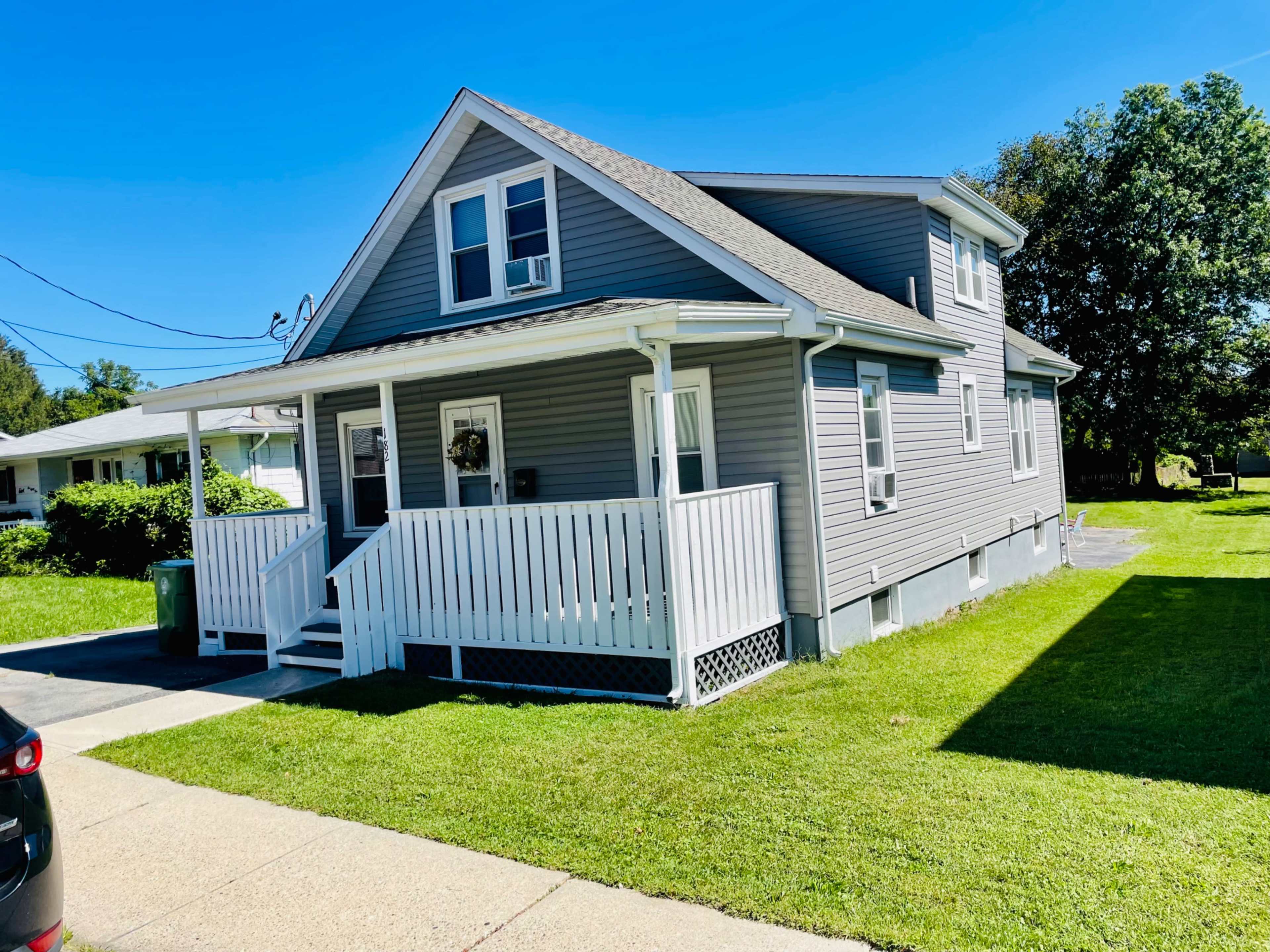 A gray two-story house with a white front porch and well-maintained green lawn is situated on a residential street.