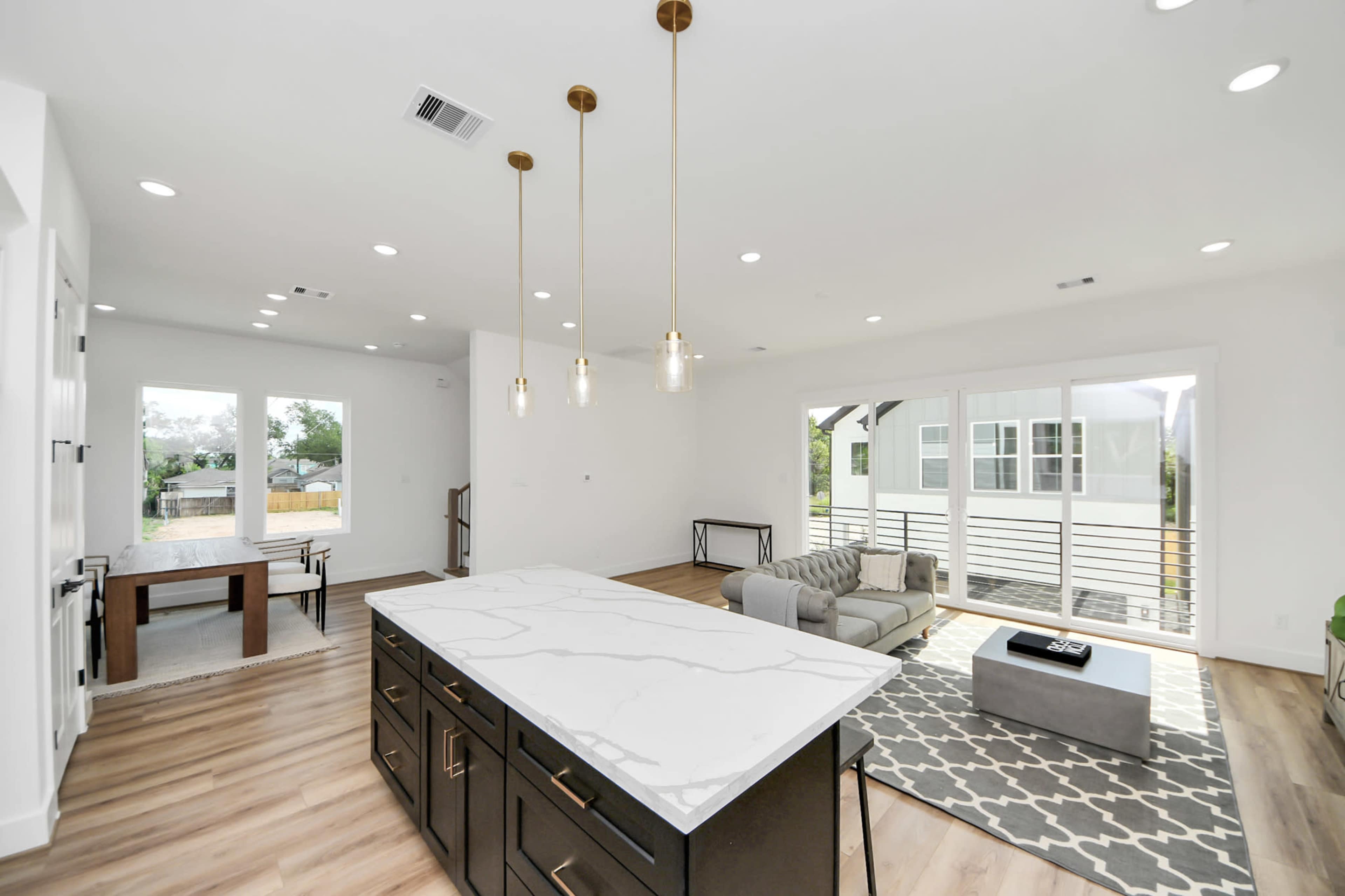 The image shows a modern open-concept living space featuring a kitchen island with marble countertops, a gray sofa, and large windows letting in natural light.