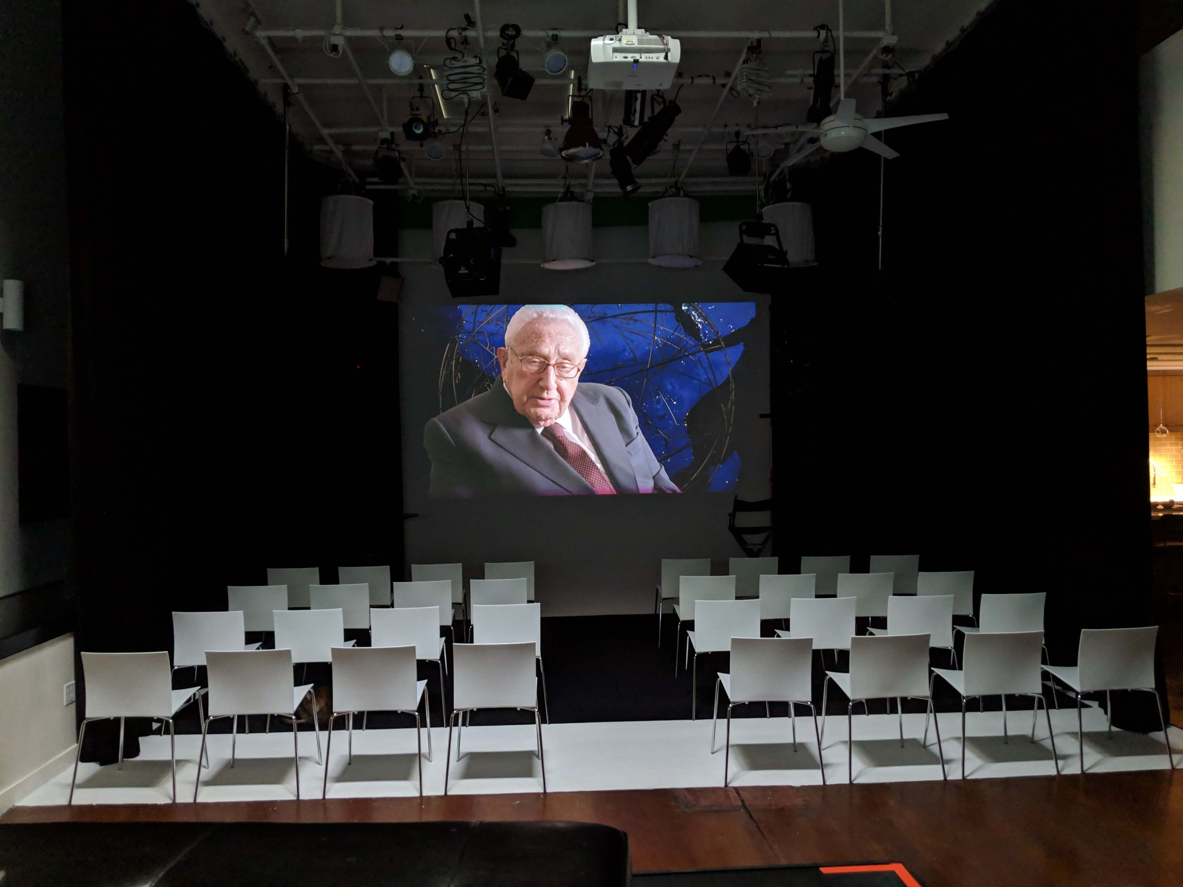 A large projection screen displays an elderly man in a suit, while empty white chairs are arranged in rows in front of it in a dimly lit room.