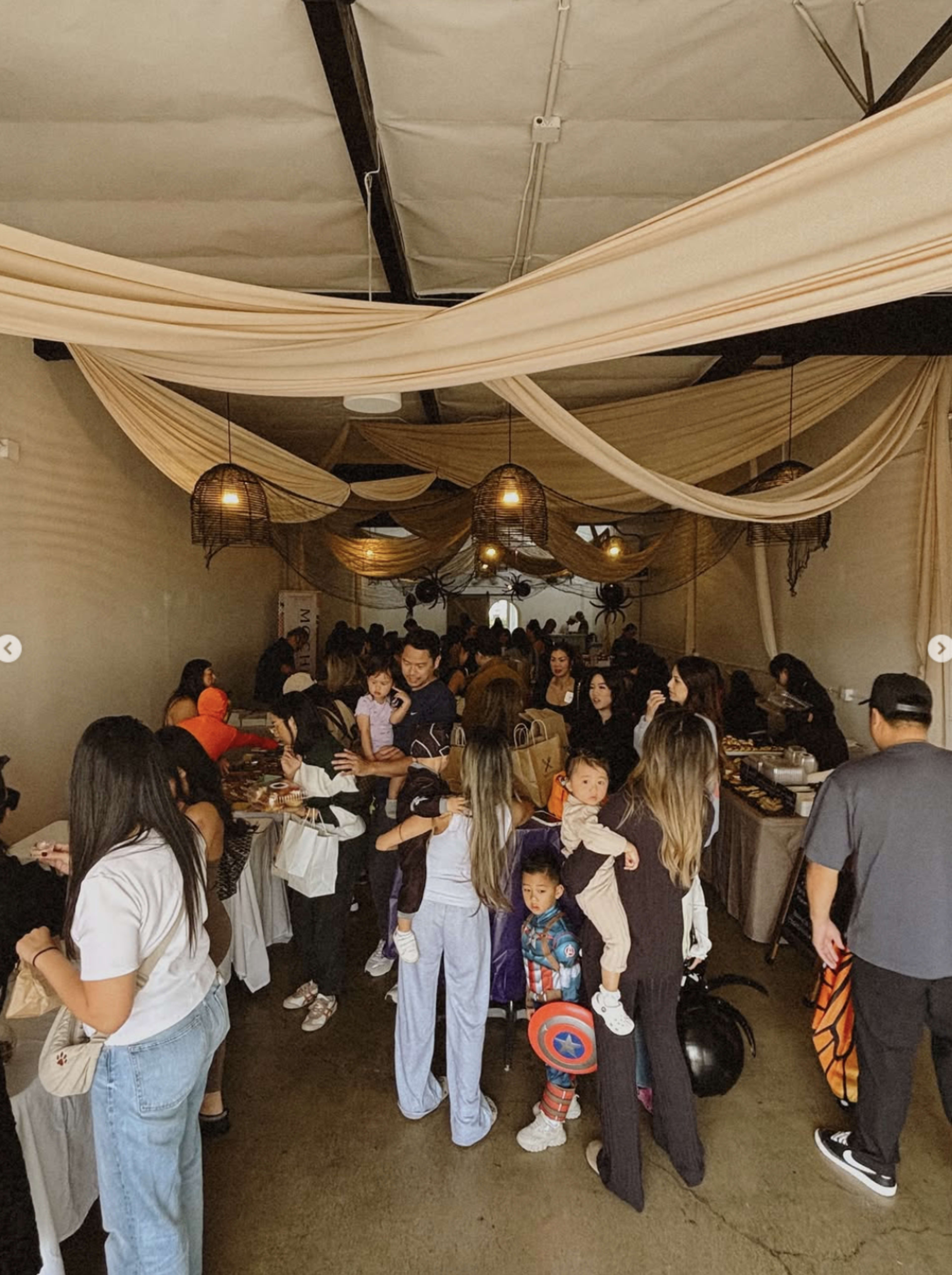 A crowded indoor space is filled with people celebrating an event, featuring tables with food and decorations, while children in costumes mingle among the adults.