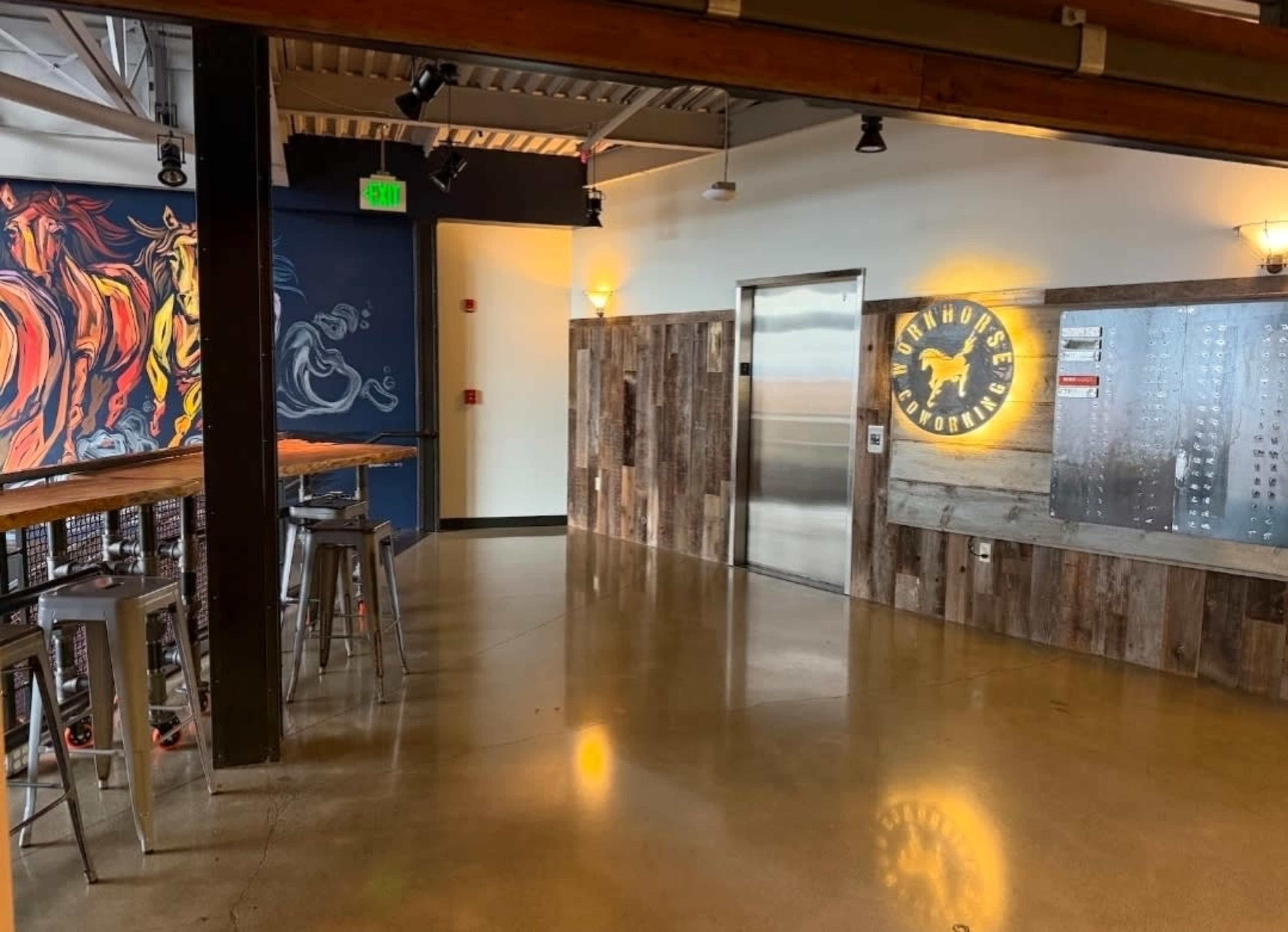 The image depicts a modern interior of a restaurant or bar featuring a rustic wooden wall, an elevator, and industrial-style seating with metallic stools.