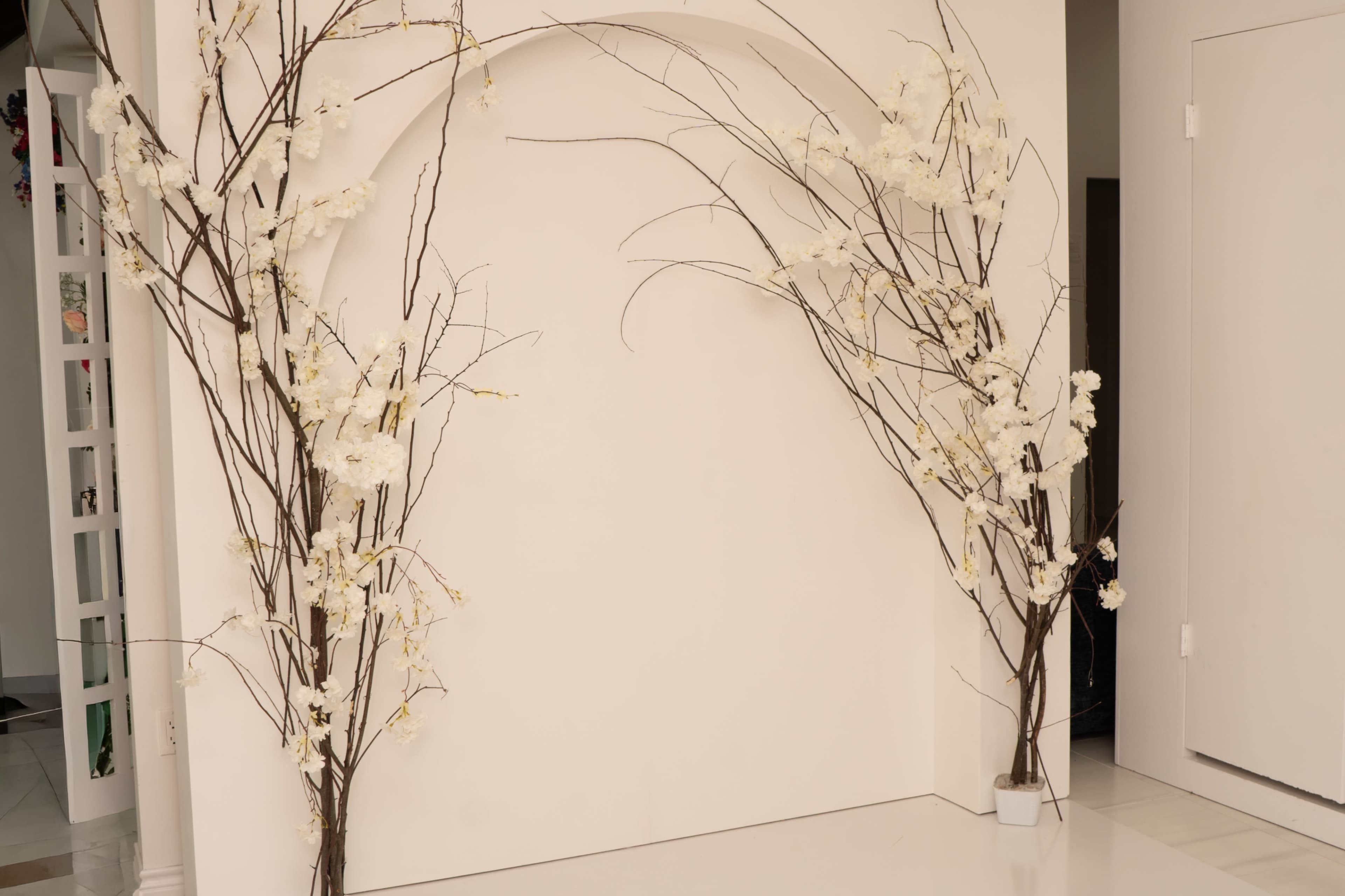 The image features a decorative arch made of bare branches adorned with white flowers, positioned against a plain white wall.